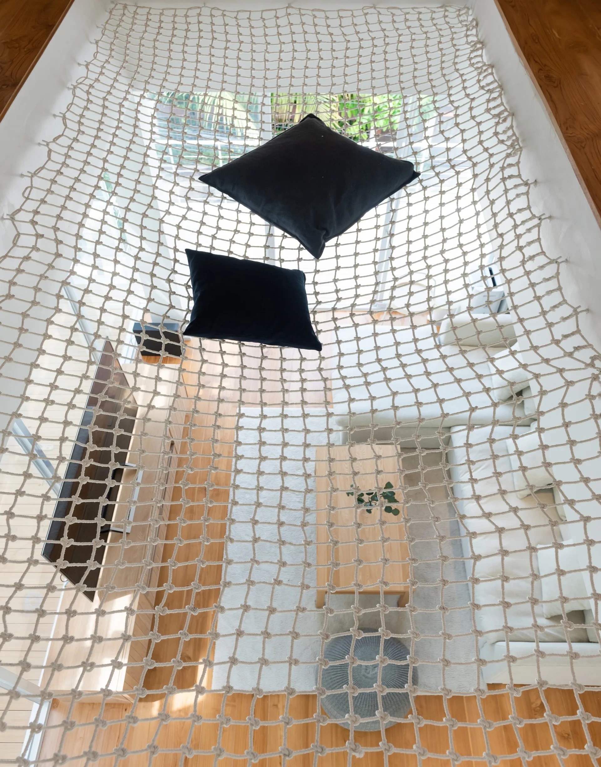 A high-angle view looking down through a woven rope net floor with black pillows into a light-filled living room with a white sofa and wood coffee table.