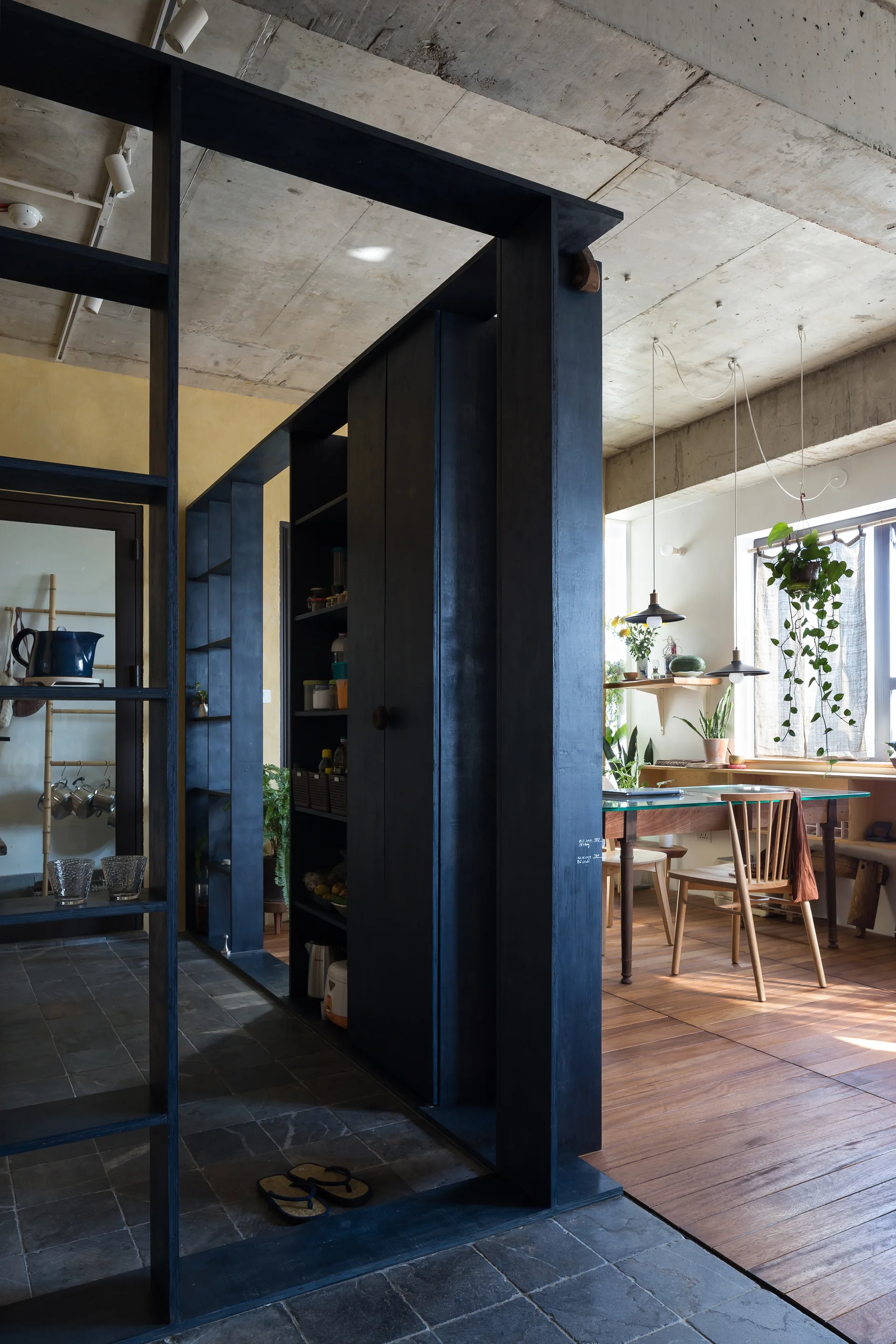 A dark wood room partition separates slate-tiled flooring from a dining area featuring wooden floors, a glass-top table, and an exposed concrete ceiling.