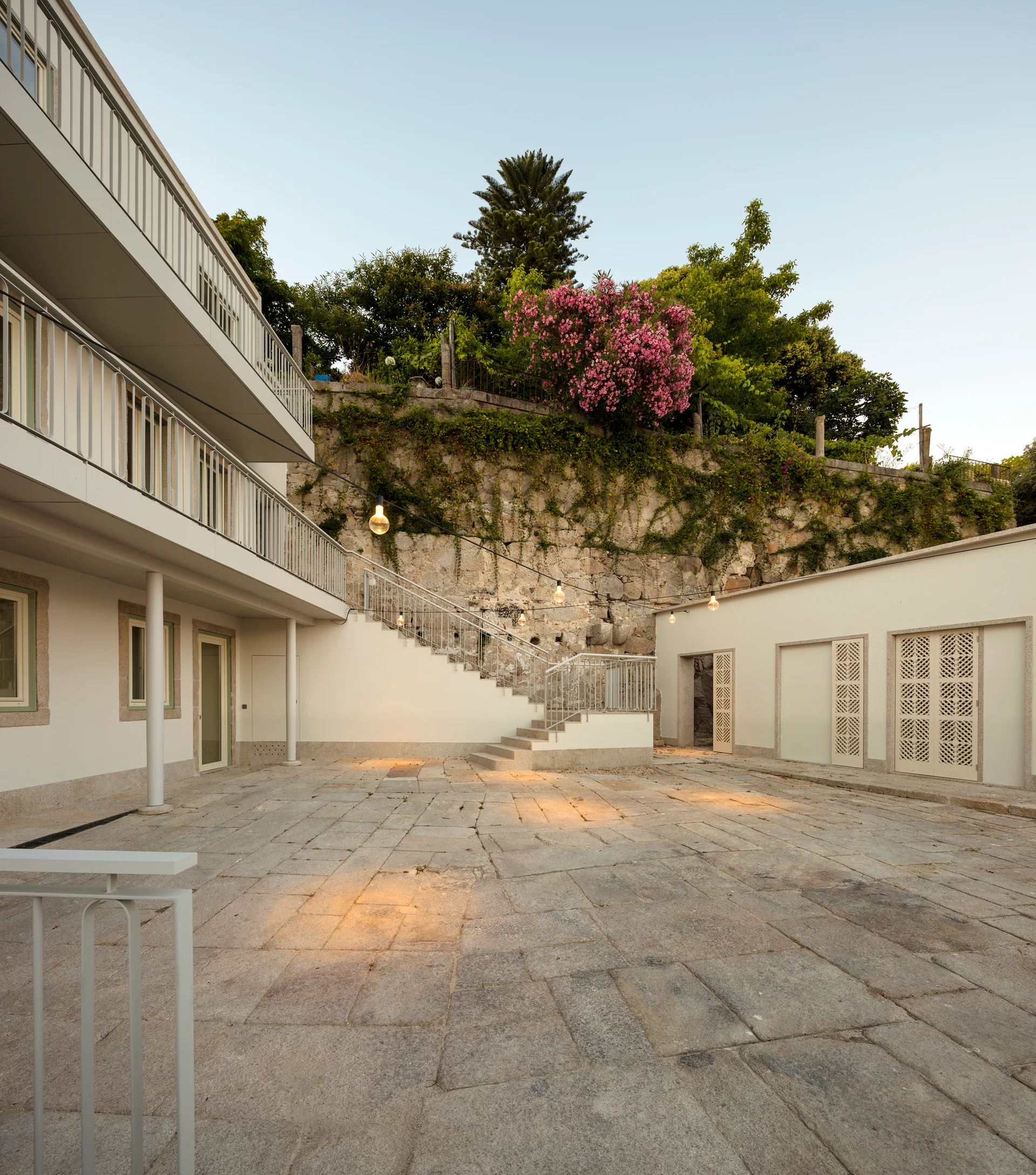 A spacious stone-paved courtyard features white building facades with balconies, an outdoor staircase, and a stone retaining wall covered in greenery.
