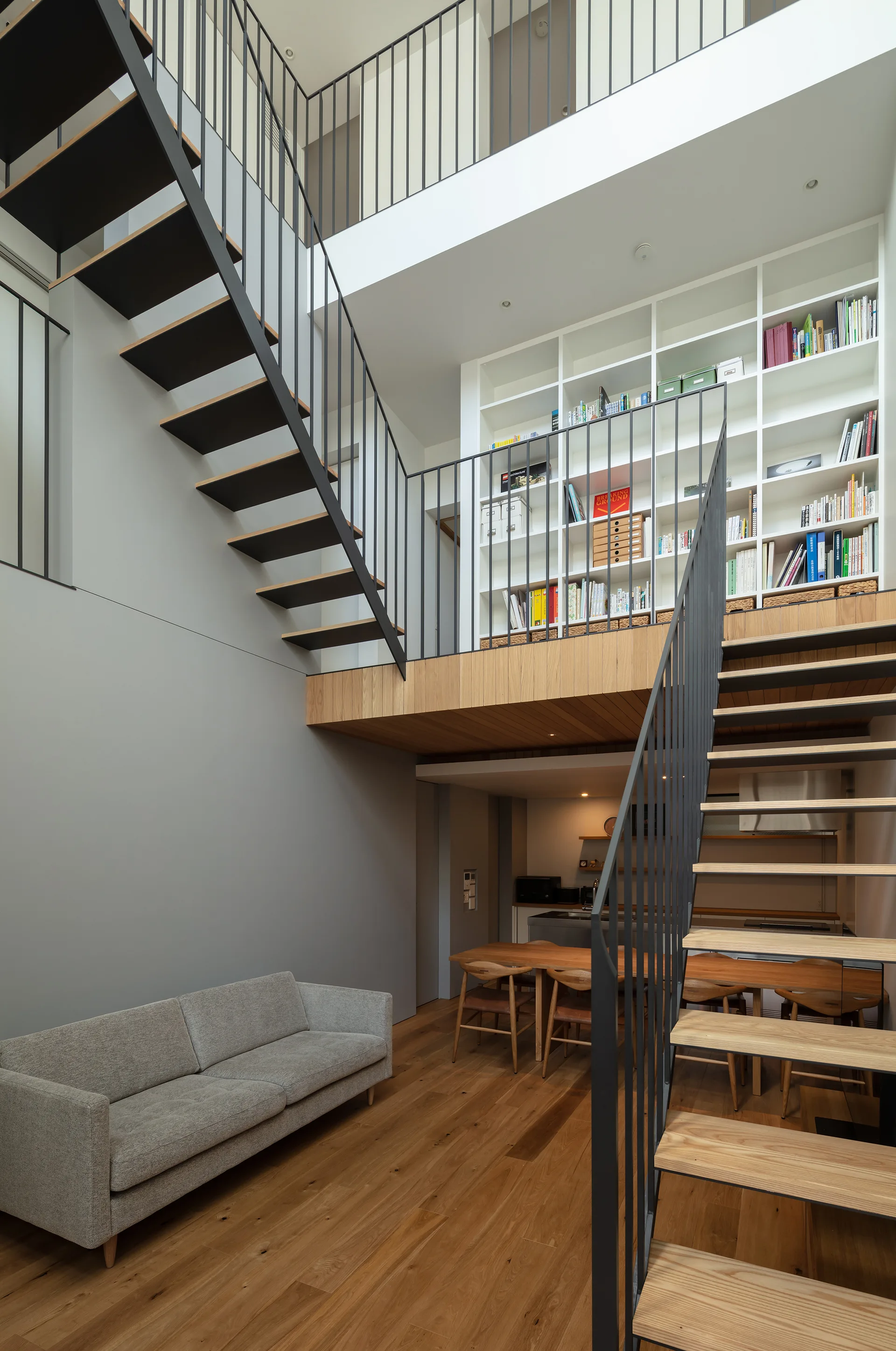 A modern open-plan living room featuring a grey sofa, wood floors, a floating staircase with black steel railings, and a mezzanine bookshelf.