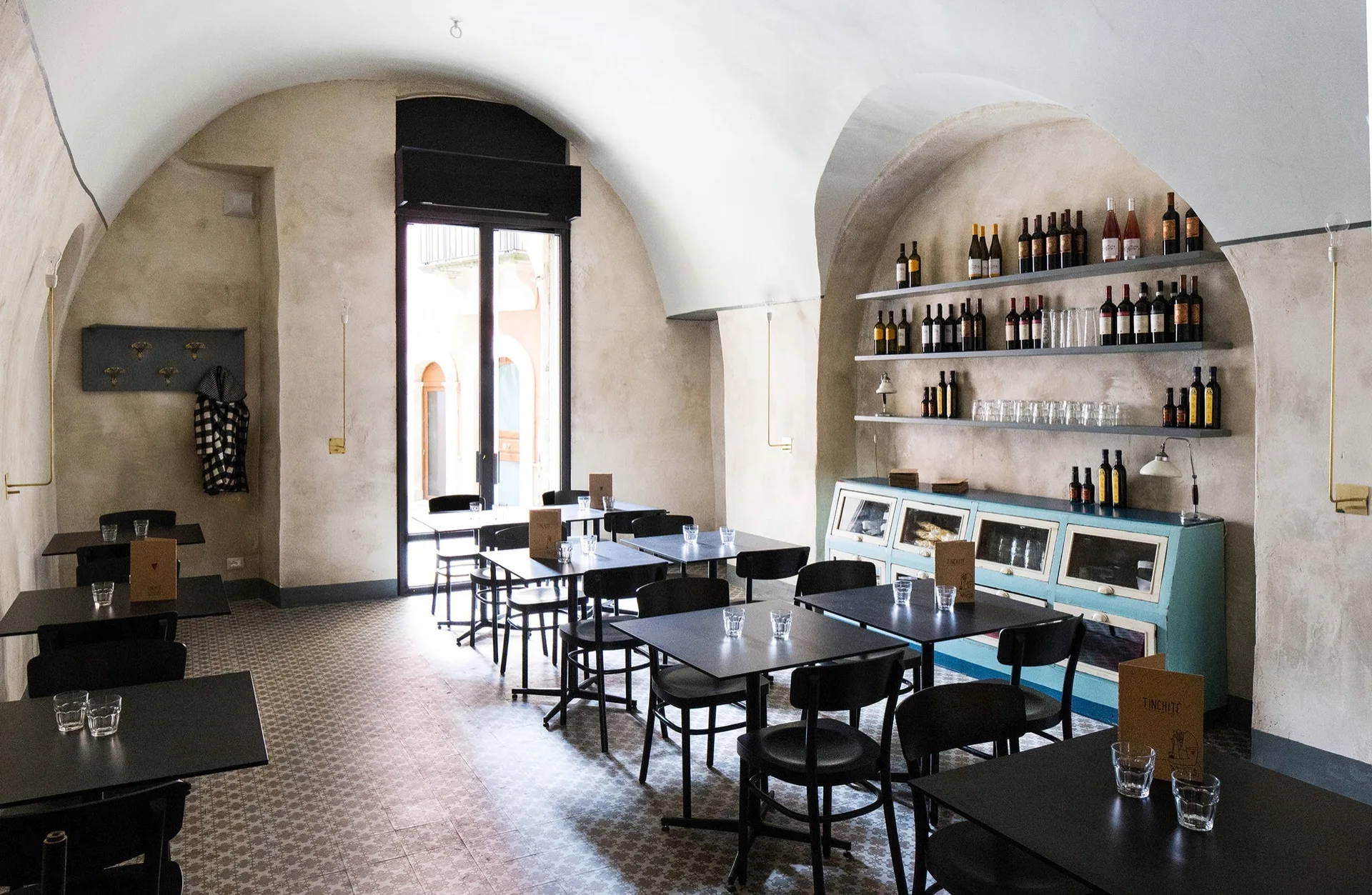 Restaurant dining room featuring vaulted plaster ceilings, black tables and chairs, geometric floor tiles, and a teal cabinet under wall shelving.