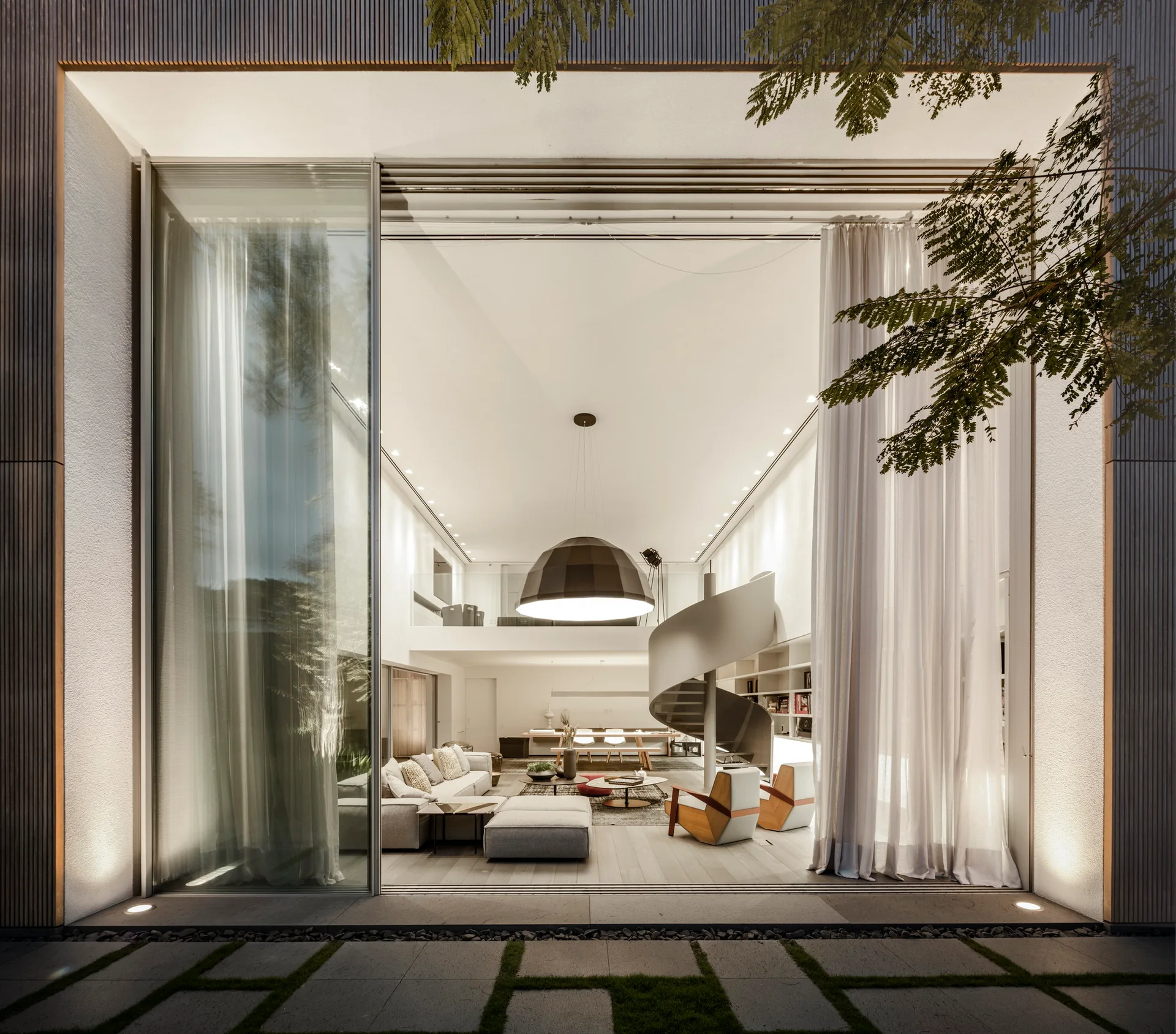 An open-concept modern living room with a metal spiral staircase, a large dome pendant light, and sliding glass walls opening to an outdoor patio.