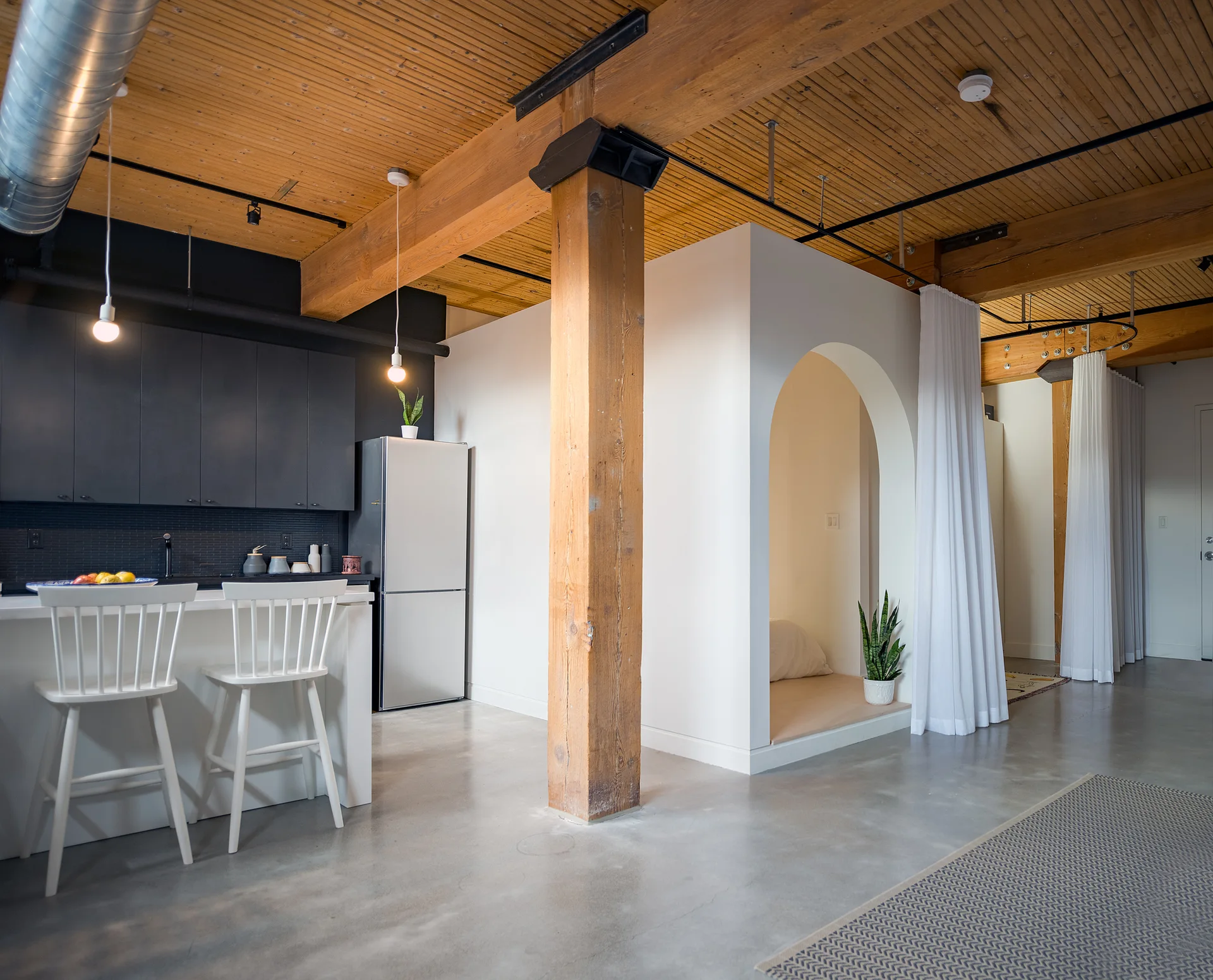 An open loft kitchen features black cabinets, a white island, concrete floors, exposed timber beams, and a white arched alcove with a privacy curtain.