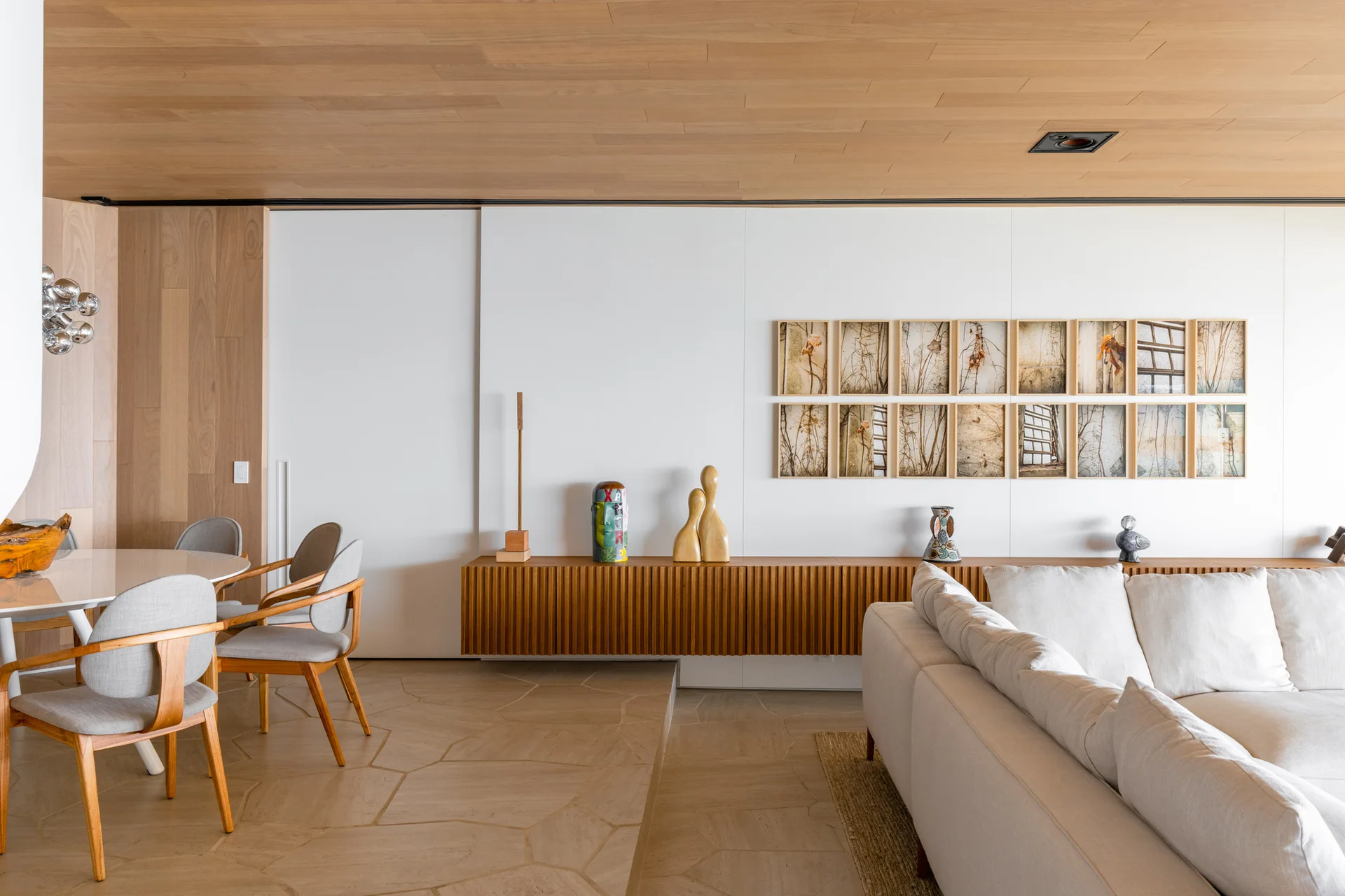 An open-plan living room features a white sofa, a wood-slatted floating console, a circular dining table, and a wood plank ceiling.