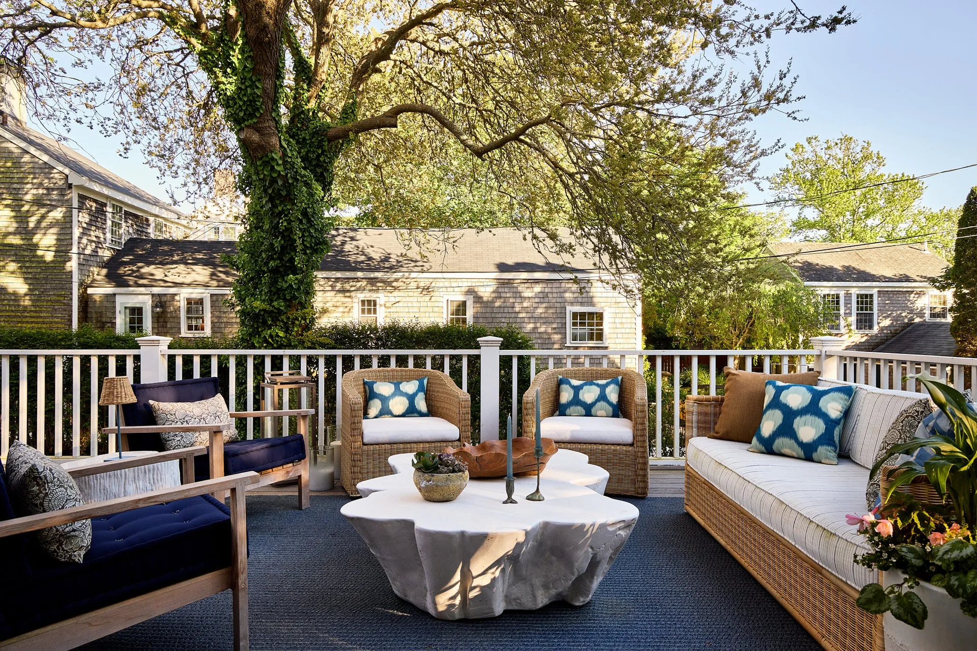 An outdoor wooden deck features a white organic-shaped coffee table surrounded by wicker and wood lounge chairs with blue cushions.