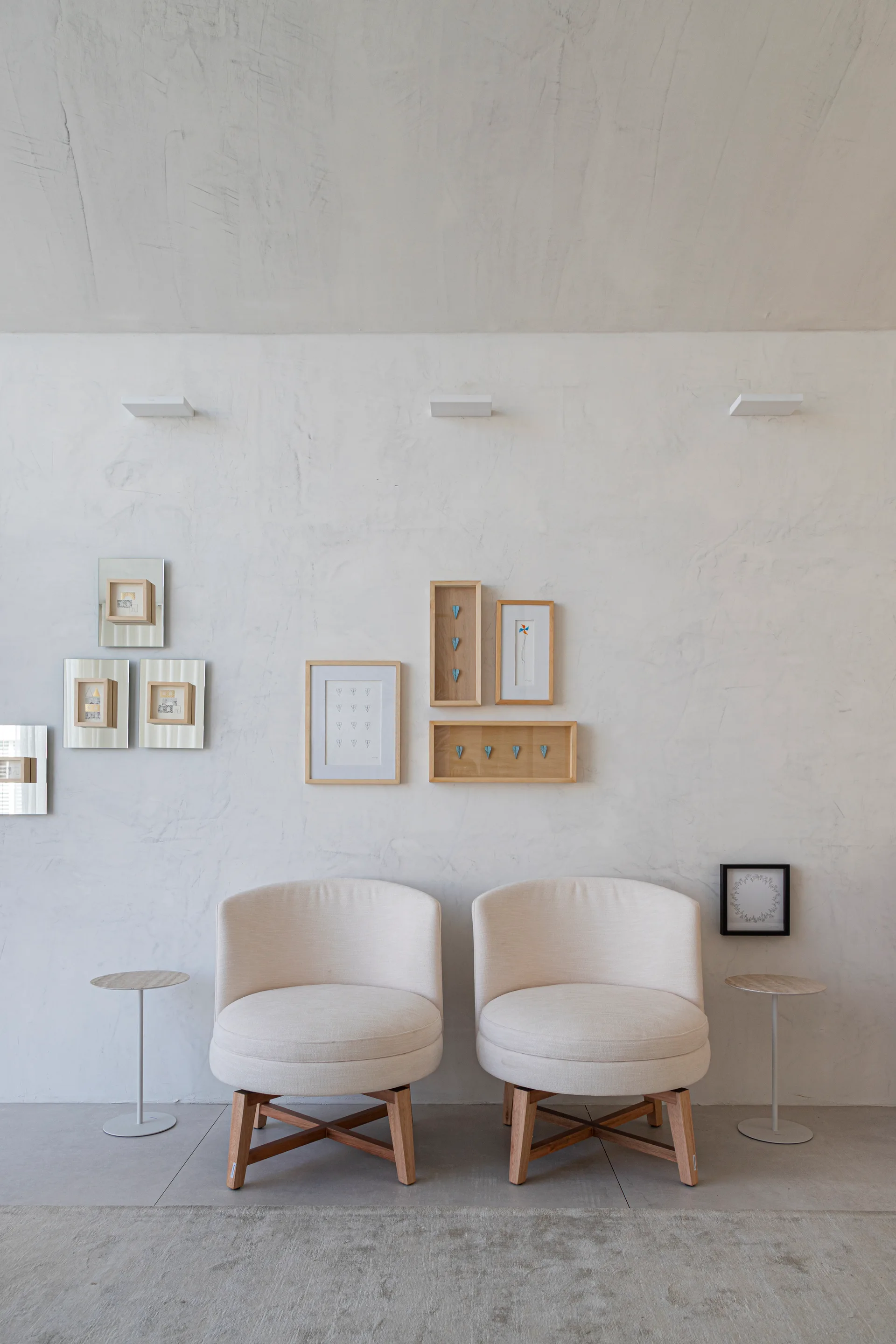 Two rounded white armchairs with wooden bases sit against a textured plaster wall decorated with framed art and small white sconces.