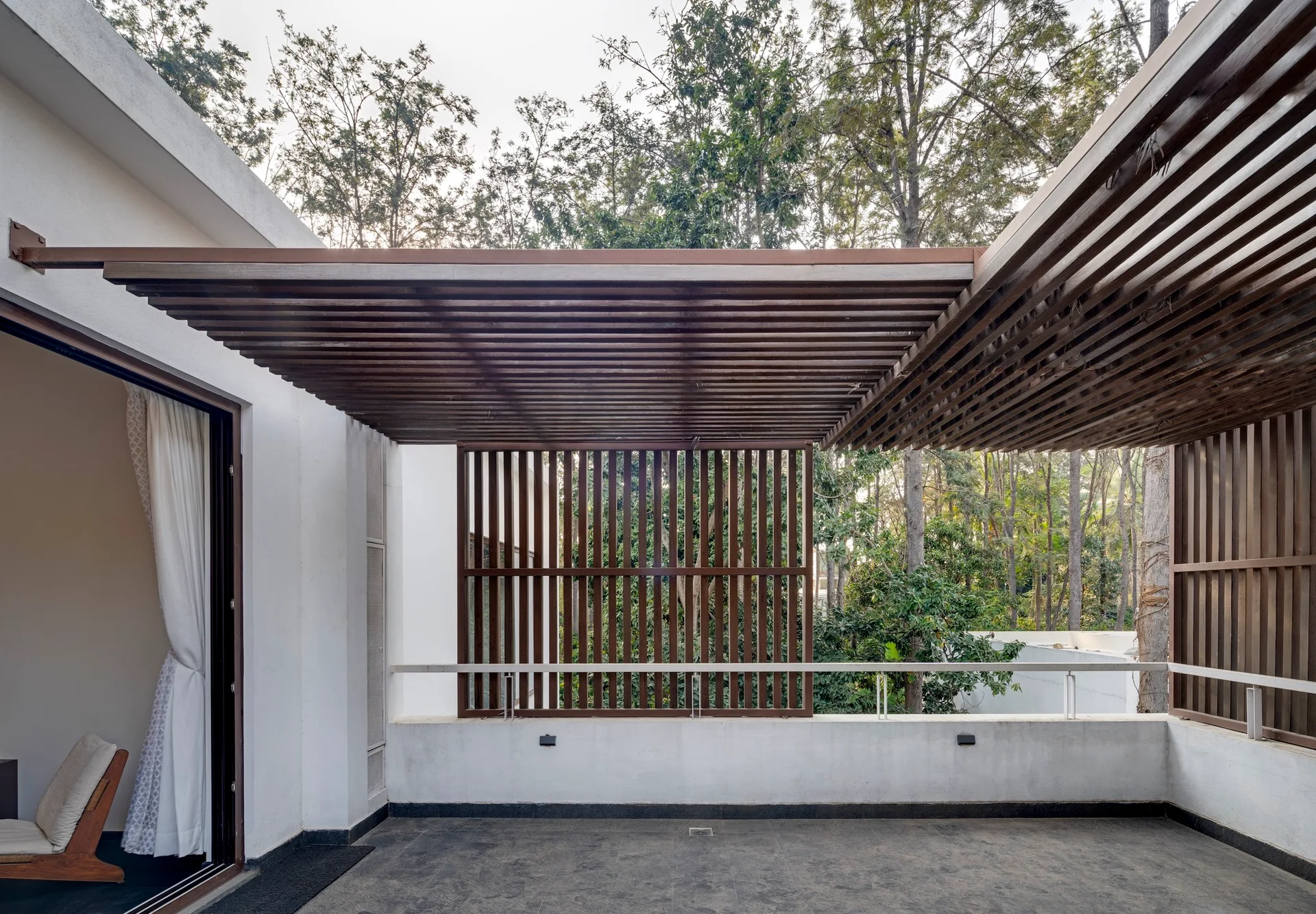 A modern outdoor terrace features a brown slatted wooden pergola overhead and a matching vertical privacy screen overlooking a forest of tall trees.