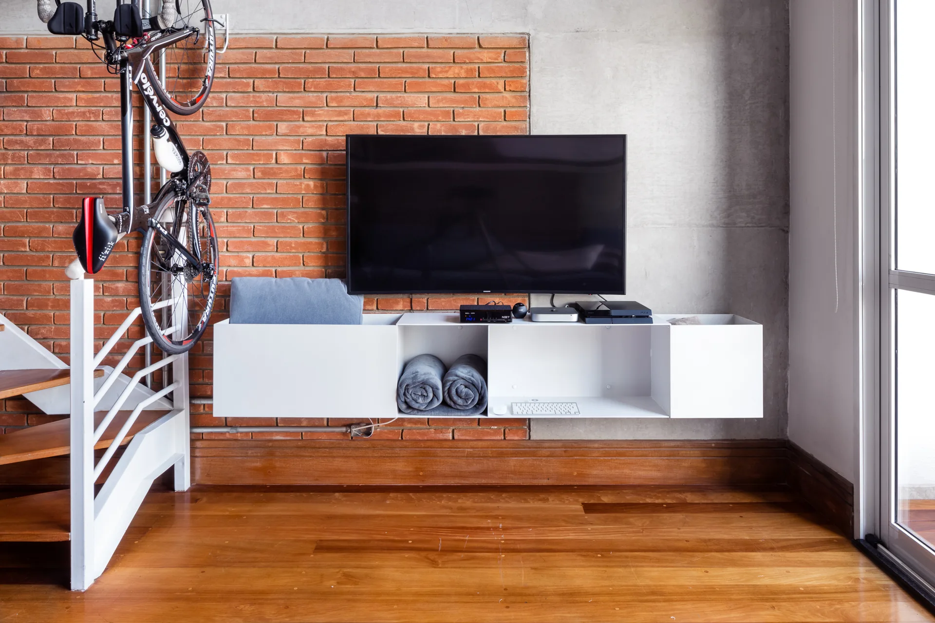 A television hangs above a white floating media console against a wall of exposed brick and gray concrete.