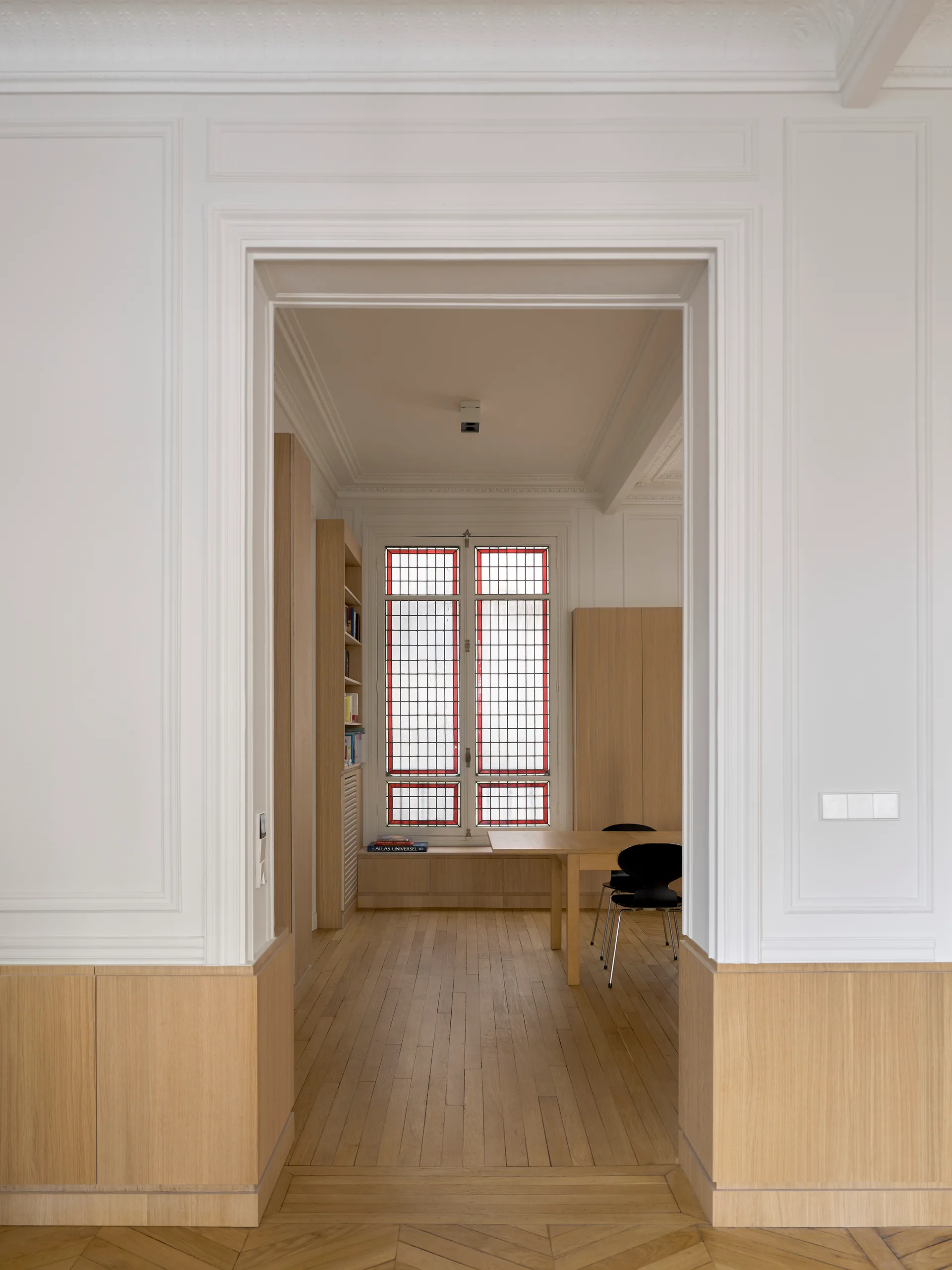 A view through a doorway into a dining area with light wood wainscoting, parquet floors, a wooden table, and a red-trimmed stained glass window.