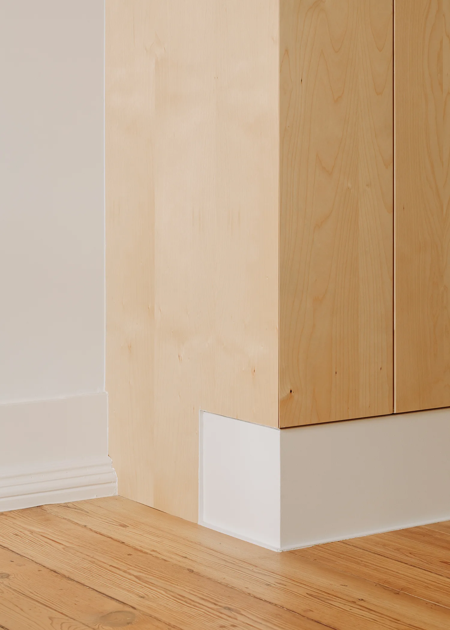 A close-up of a light wood cabinet base intersecting with a white painted wall, white baseboard trim, and natural wood plank flooring.