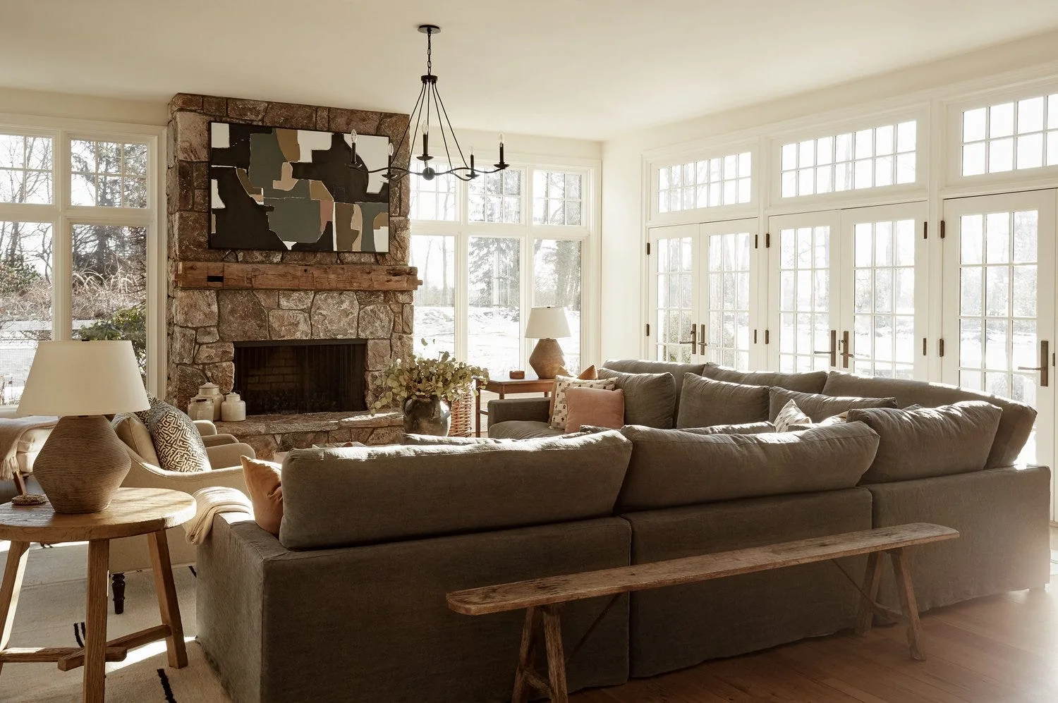 A bright living room features a stone fireplace with a rustic mantel, a large grey sectional sofa, and a slim wooden bench on wood floors.