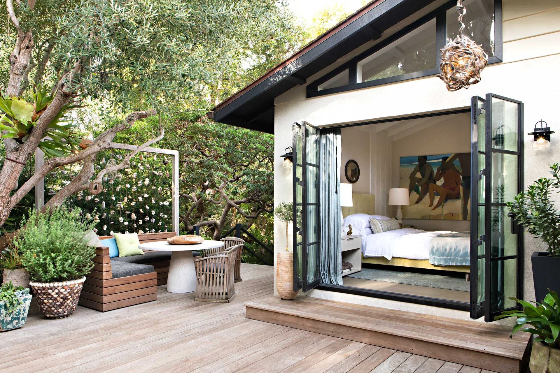 A wooden deck with built-in bench seating transitions through open black bi-fold doors into a bright bedroom with a vaulted ceiling.