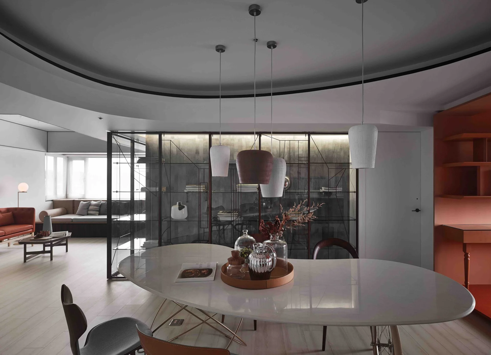 A modern dining space features an oval white table below hanging pendant lights, a circular recessed ceiling, and a glass partition divider.
