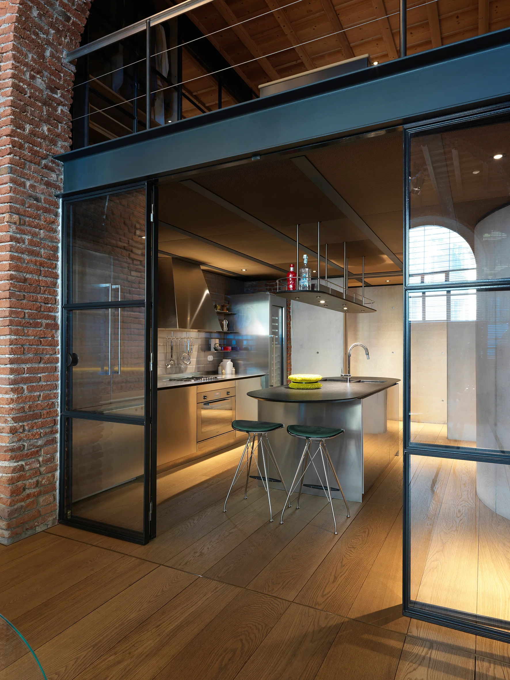An industrial kitchen with a stainless steel island and bar stools, enclosed by black metal-framed glass sliding doors next to an exposed brick wall.