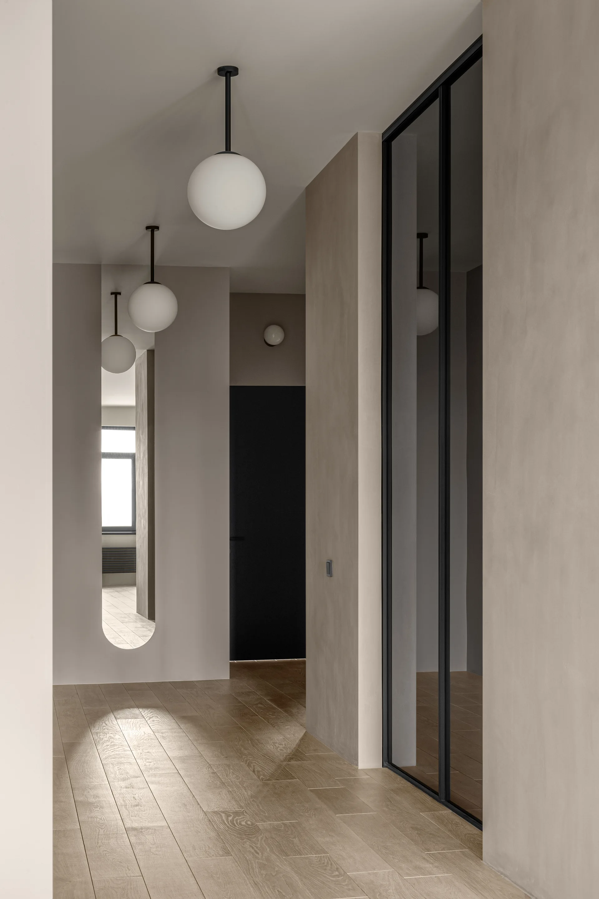 A minimalist hallway with smooth beige walls, light wood flooring, hanging white globe pendant lights, and a tall recessed wall mirror.