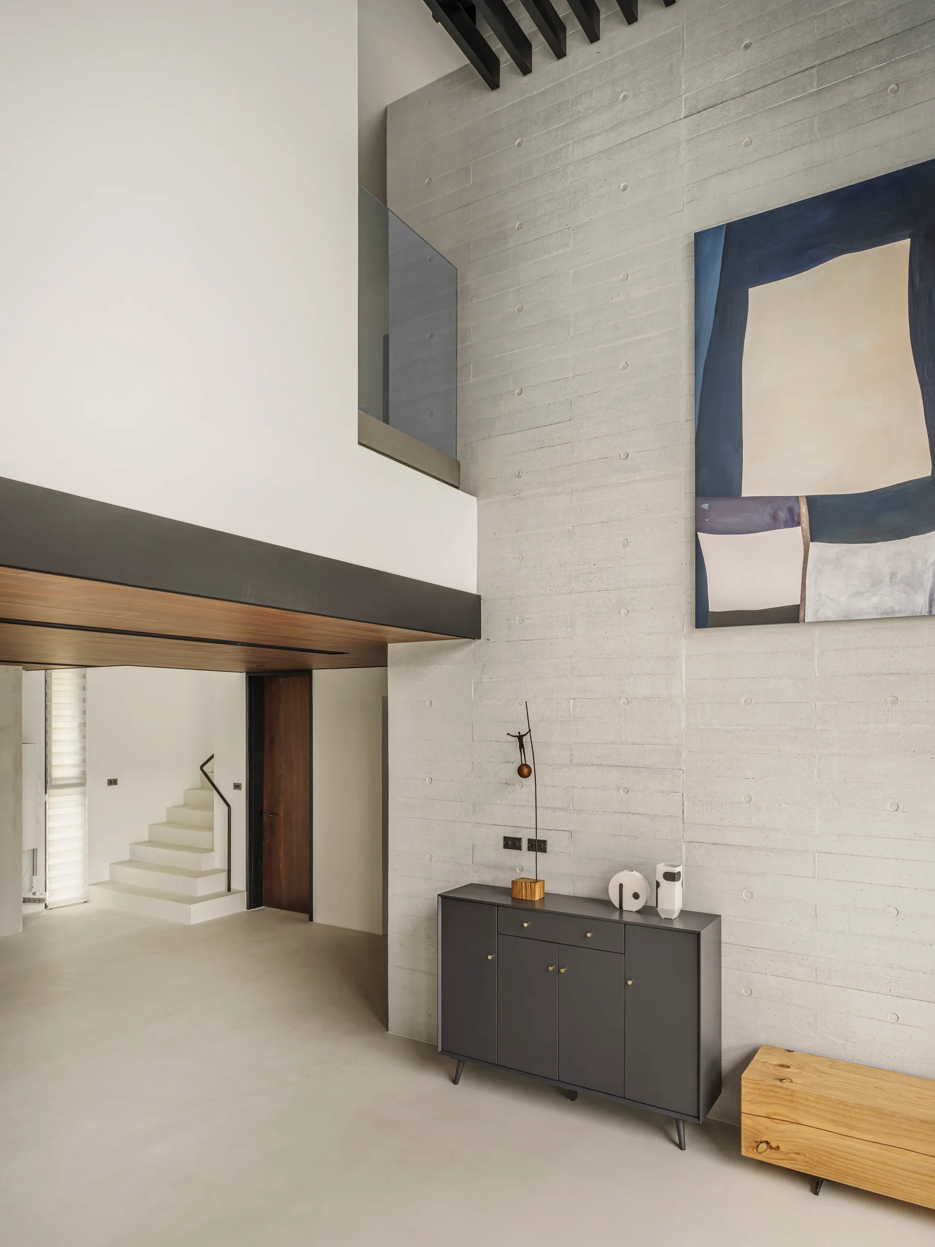 A minimalist double-height room features a textured concrete wall with an abstract blue painting, a dark grey cabinet, and a view of a white staircase.