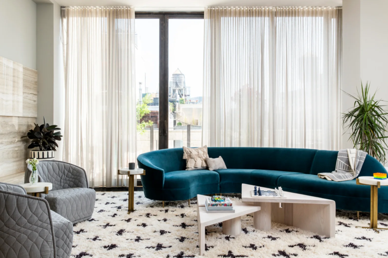 A modern living room features a curved teal velvet sofa, two gray quilted armchairs, and a tiered light wood coffee table on a patterned shag rug.