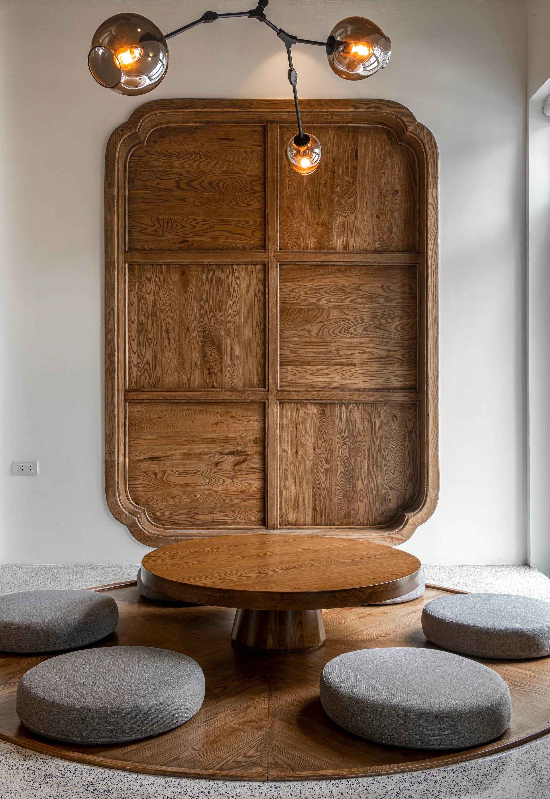 A low round wooden table is surrounded by grey floor cushions on a circular wood platform against a large matching wood wall panel.