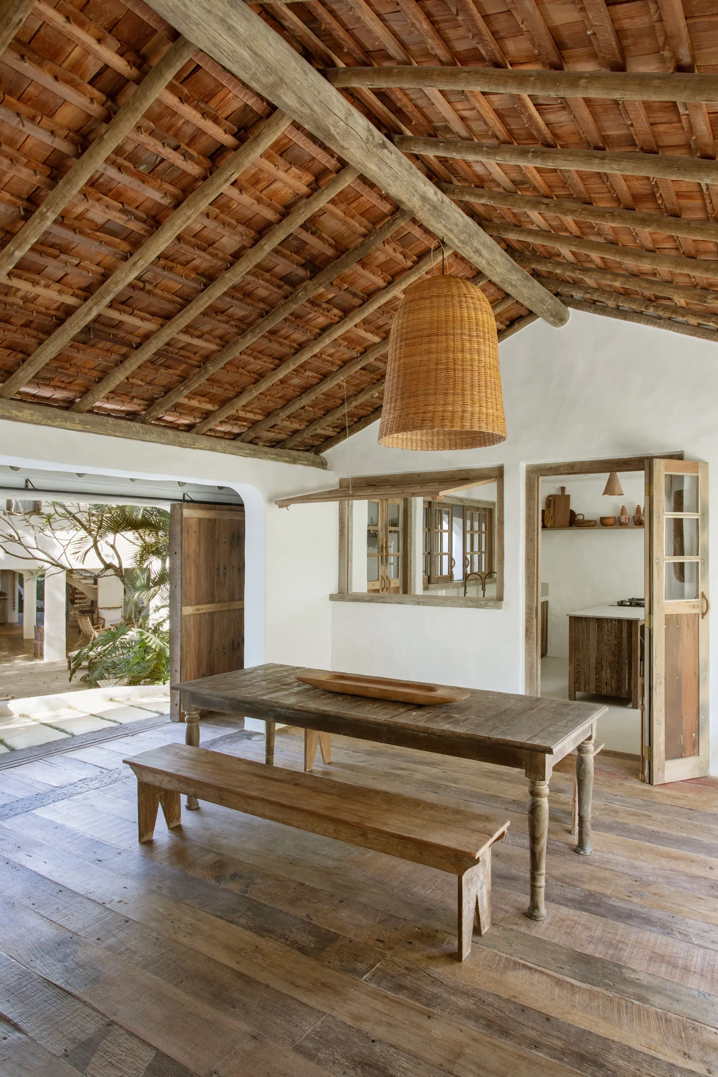 A rustic dining room features a wooden table and bench beneath a vaulted ceiling with exposed wood rafters and a large woven pendant light.