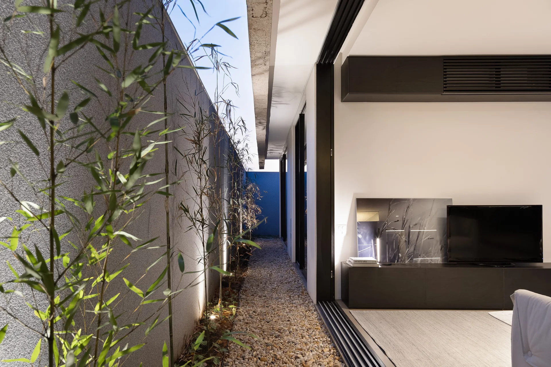 A modern living room transitions through a sliding glass door to a narrow gravel courtyard with tall bamboo plants and outdoor up-lighting.