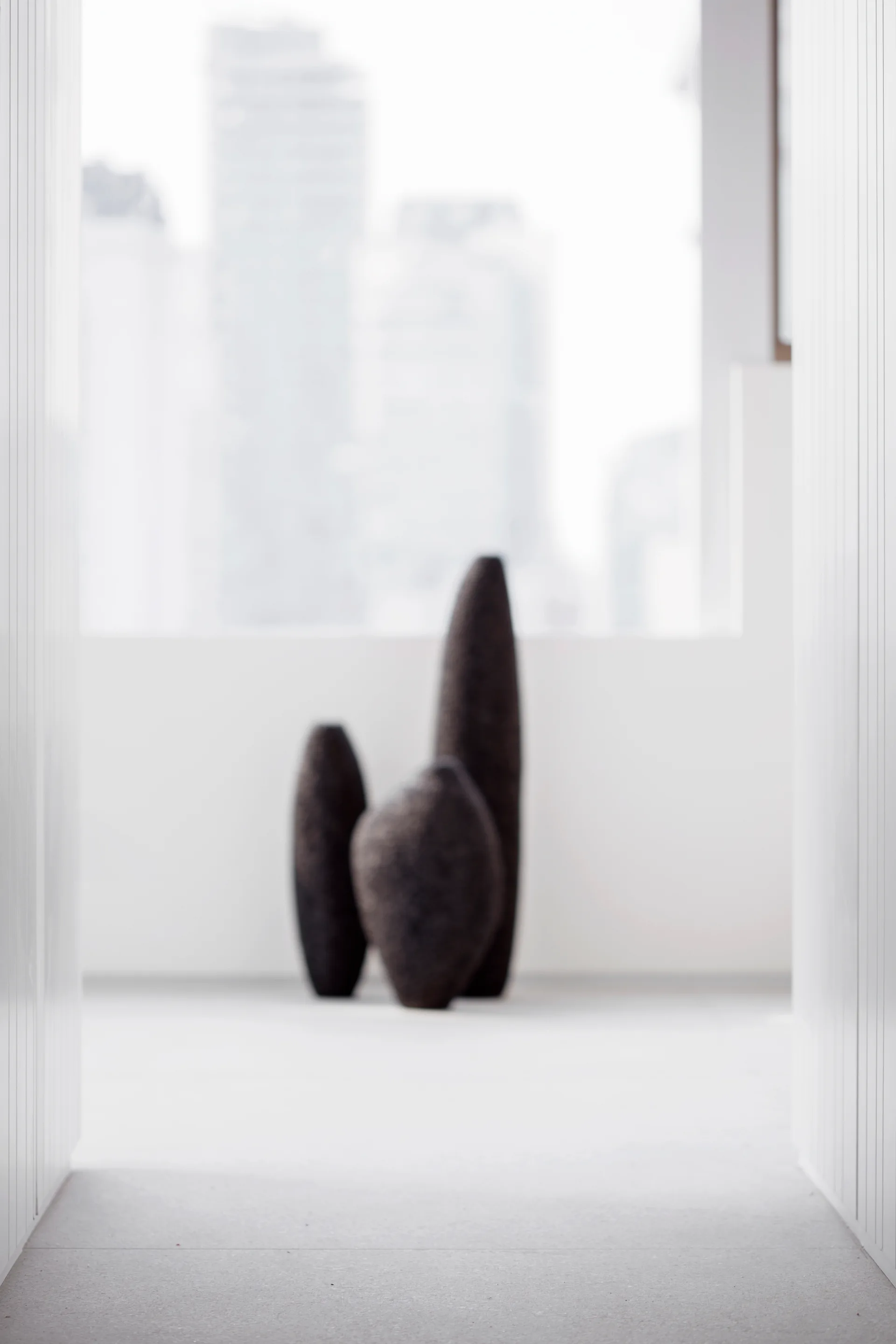 Three tall, dark, textured organic sculptures stand in a bright, minimalist white room with a blurred window in the background.