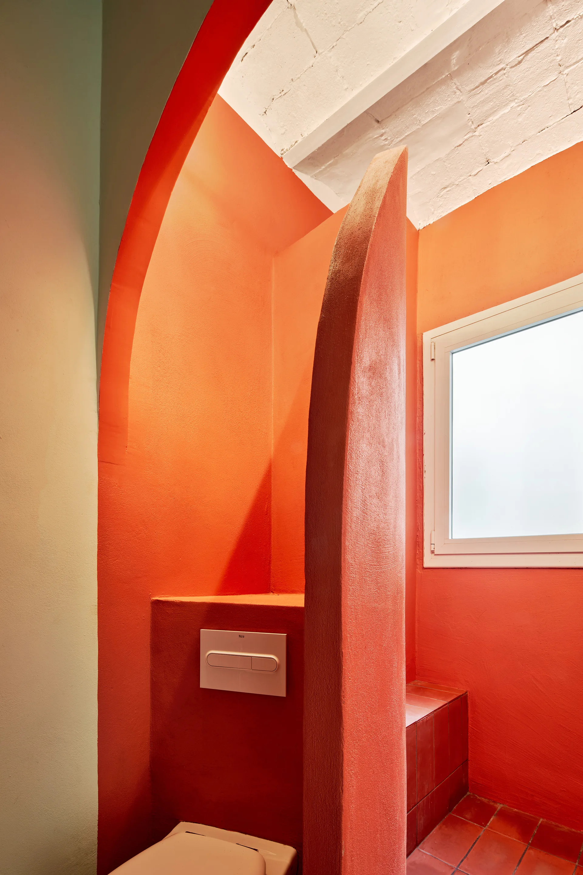 An orange powder room features a curved plaster partition wall, terracotta floor tiles, and a white brick ceiling.