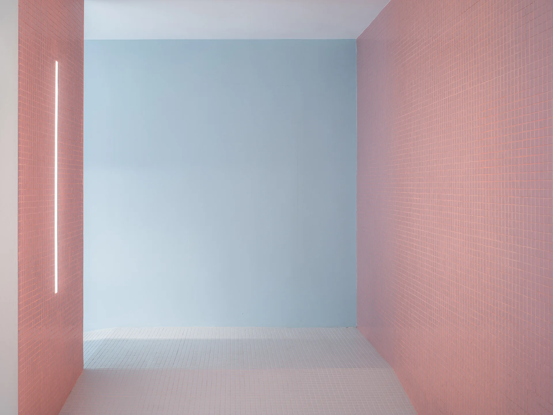 A minimalist corridor features pink grid-tiled walls, a smooth light blue accent wall, a white tiled floor, and a vertical linear light strip.