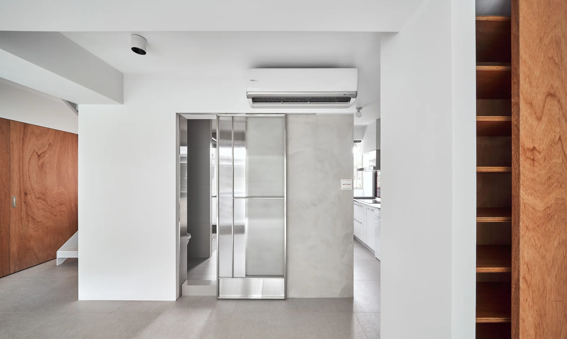 A minimalist hallway features a frosted glass sliding door with a metal frame, concrete-finish wall, and natural wood built-in shelving.