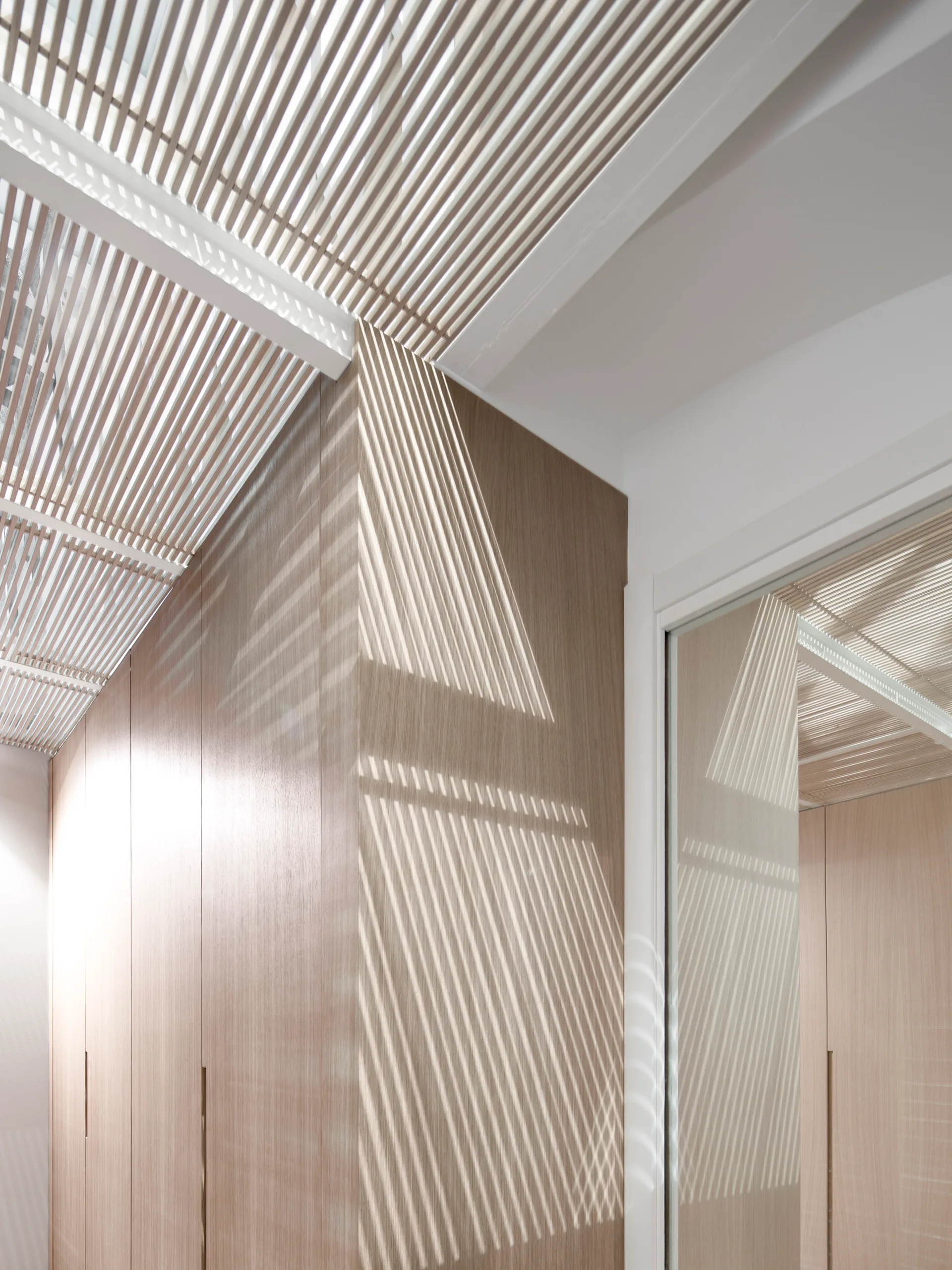 A white slatted ceiling casts linear shadows across minimal, light-wood panel cabinetry in a contemporary hallway.