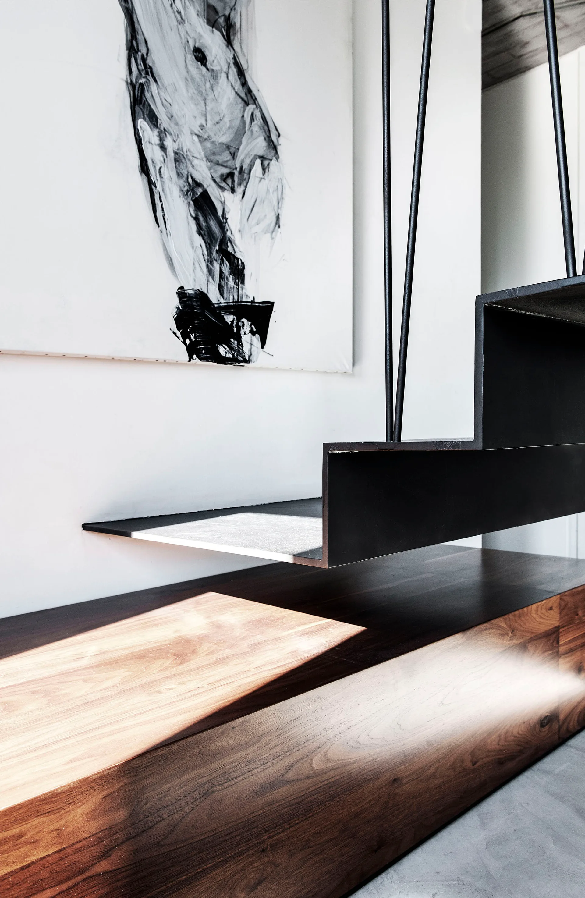 A close-up of a black metal cantilevered stair tread hovering above a low wood cabinet against a white wall with abstract art.