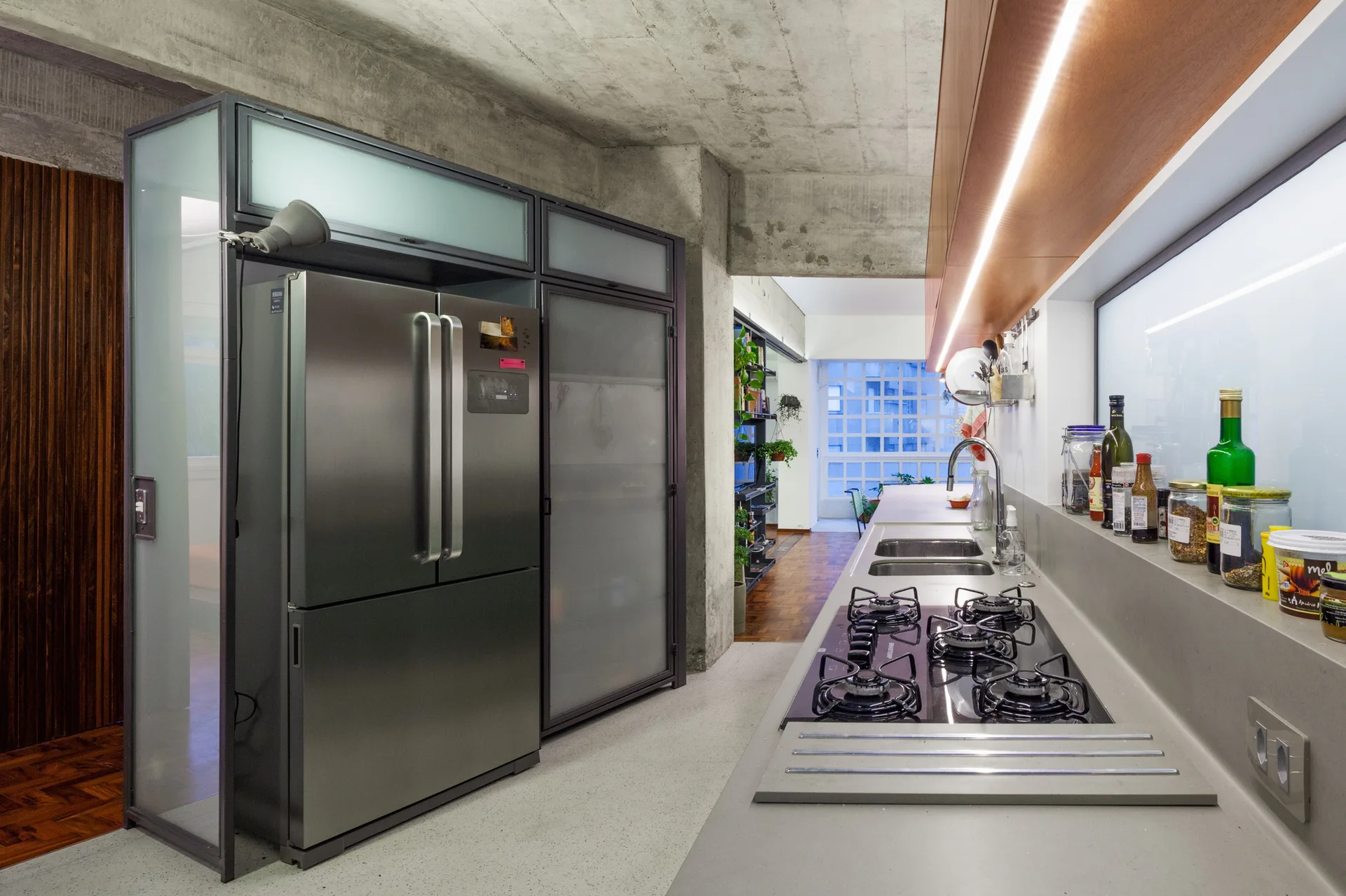A modern kitchen featuring a stainless steel refrigerator in a metal-framed cabinet, concrete ceiling, and a grey countertop with a gas cooktop.