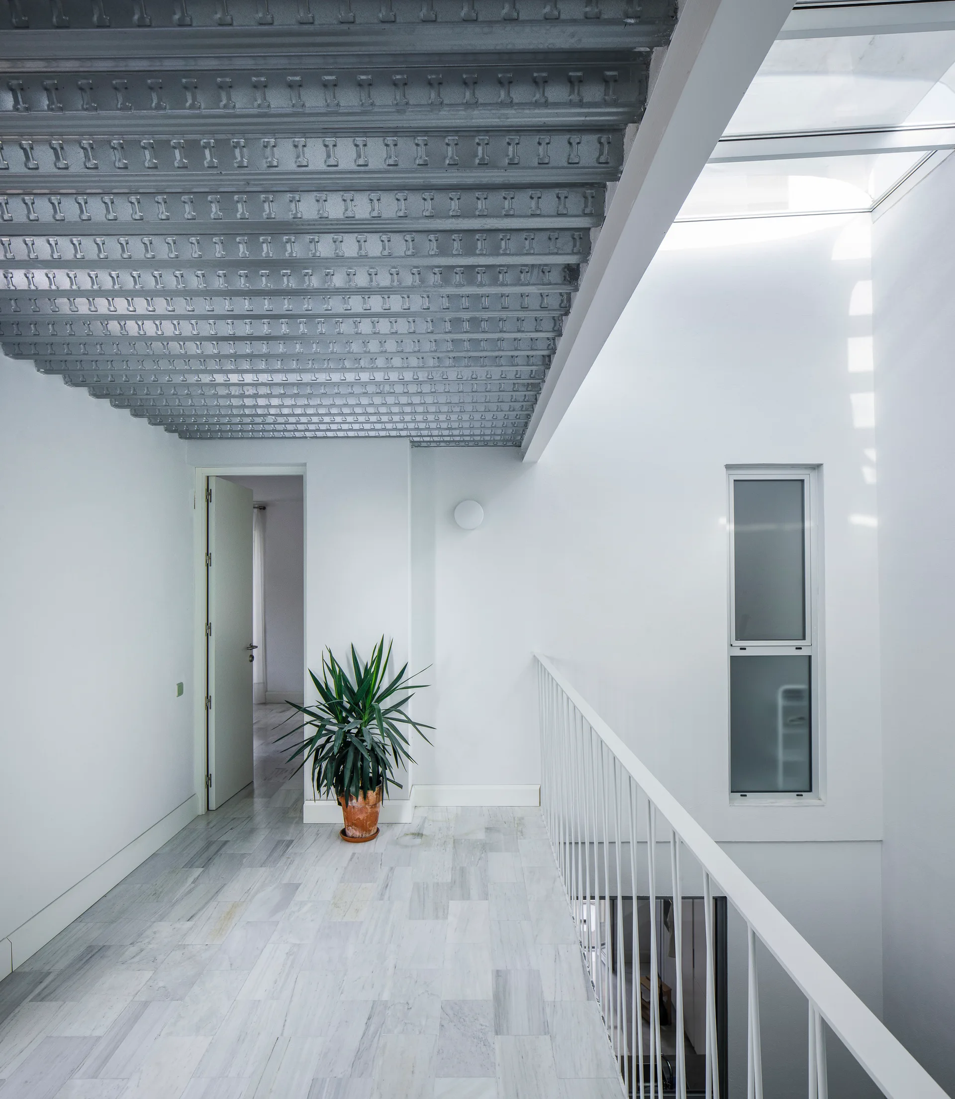 A bright white hallway landing features an exposed corrugated metal ceiling, veined marble floors, a white metal railing, and a tall green potted plant.