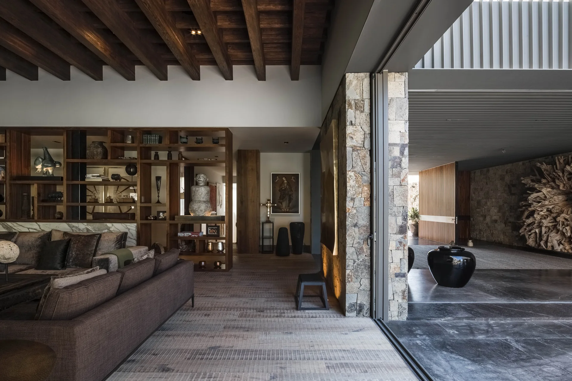 A modern living room features a dark brown sofa, exposed wood ceiling beams, a stone wall column, and a large sliding glass door opening to the outdoors.