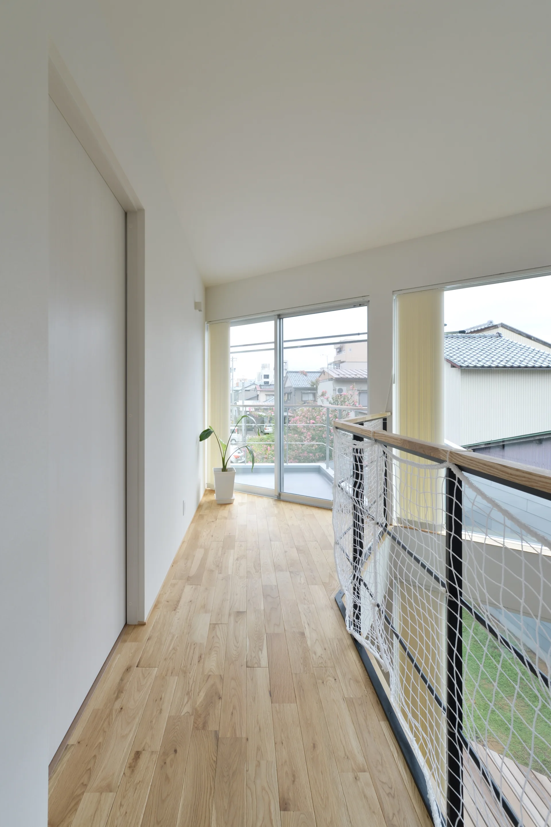 A bright hallway landing features light wood flooring, white walls, and a black metal railing with white safety netting next to a glass door.