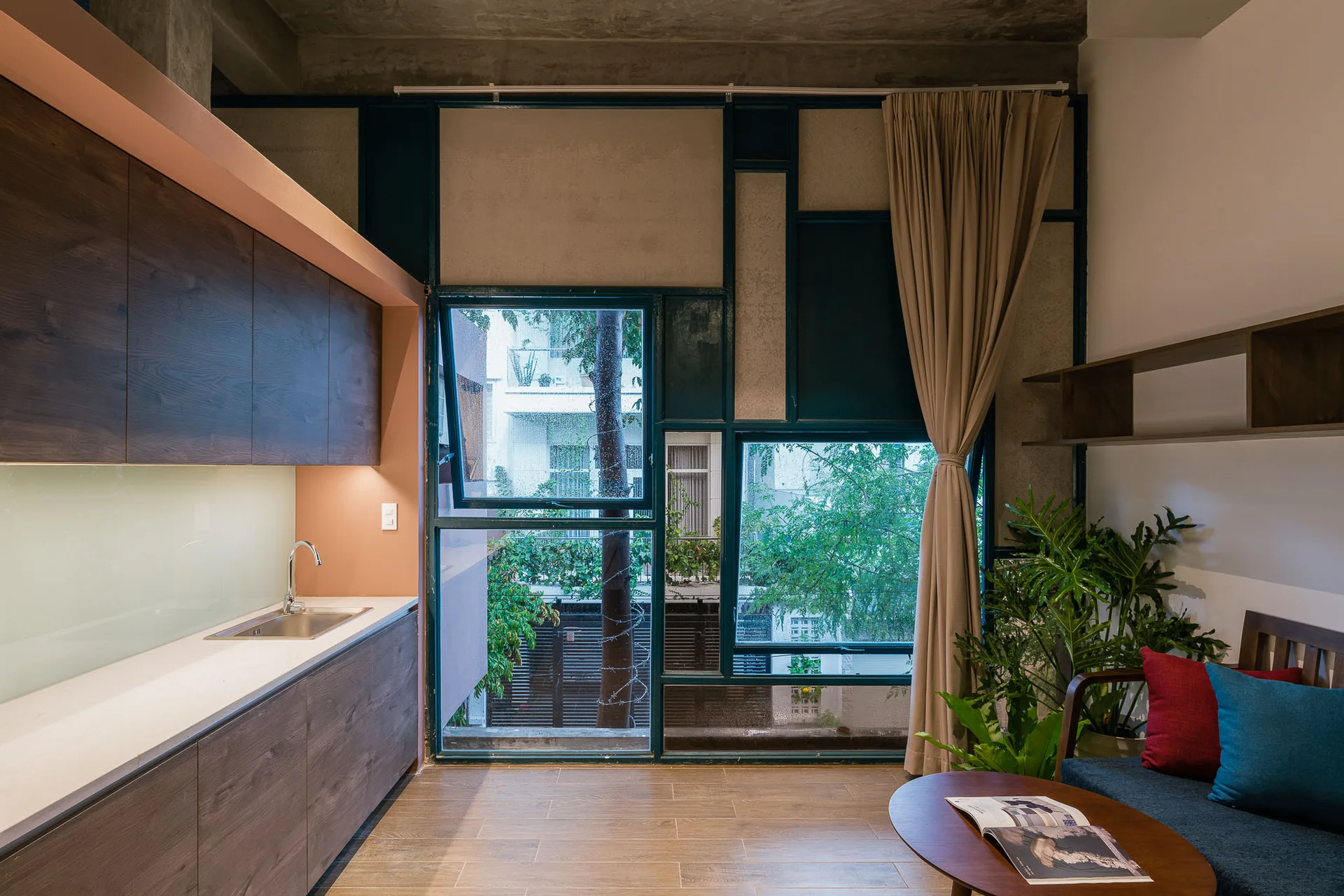 A modern kitchenette with dark wood cabinetry features a large geometric metal window and a concrete ceiling next to a small seating area.