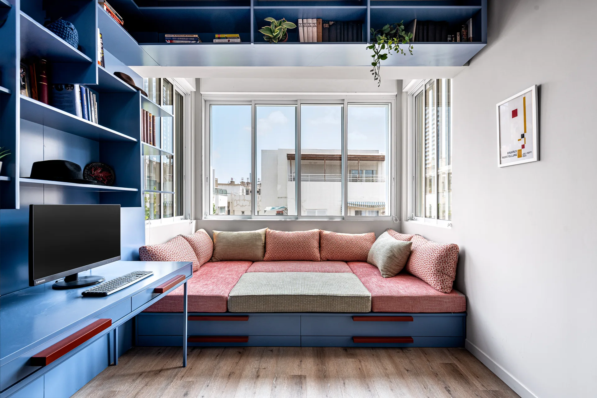 A home office featuring custom blue built-in cabinetry with an integrated desk, a window daybed with coral cushions, and light wood floors.