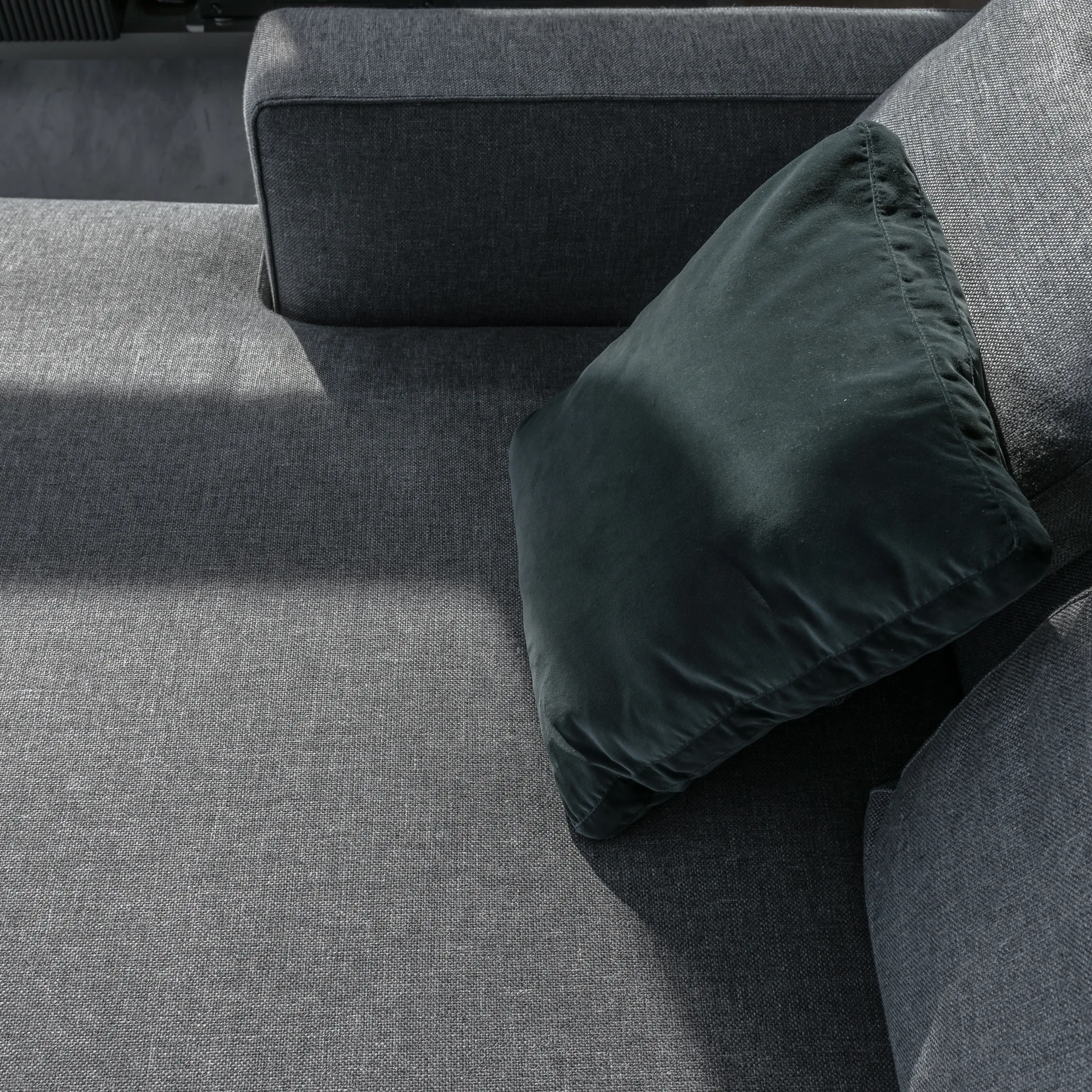 A close-up view of a textured grey fabric sofa with a square dark green velvet throw pillow resting on it in natural sunlight.