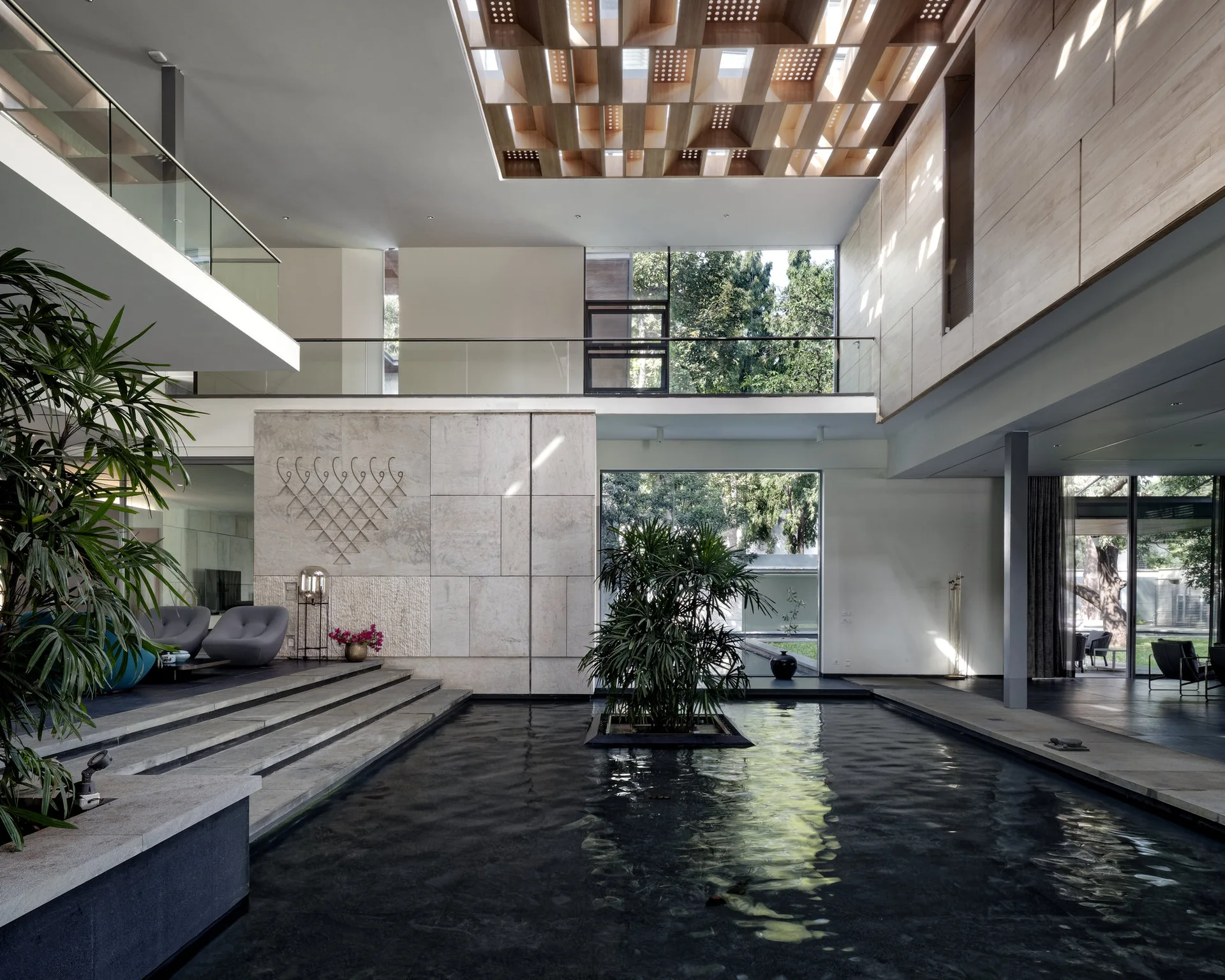 A spacious modern interior atrium featuring a large indoor water pool, a geometric wooden grid skylight, stone walls, and a glass-railed balcony.