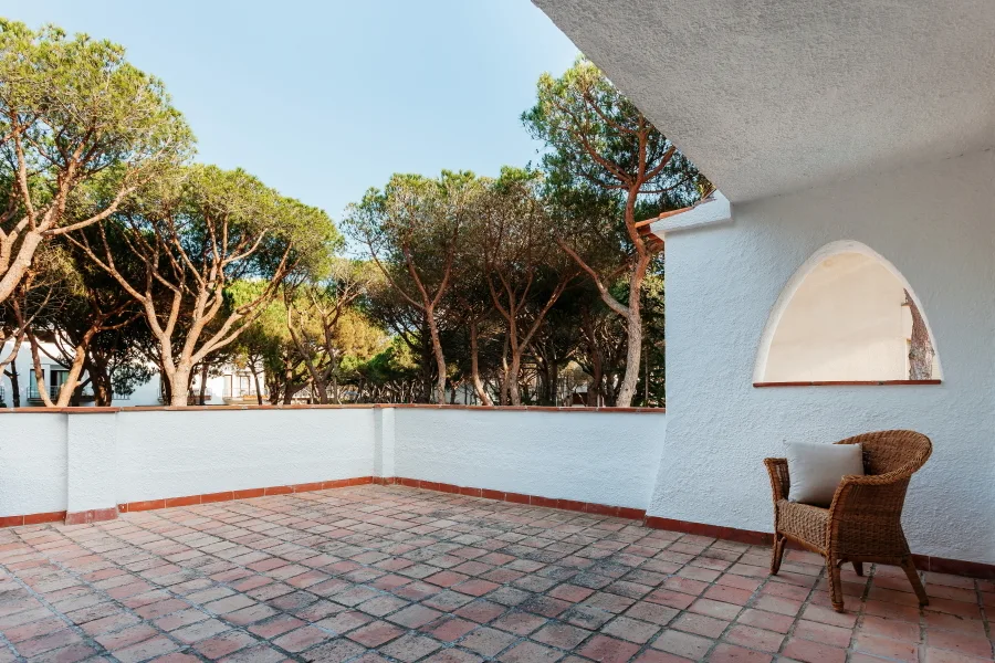 An outdoor terrace with terracotta floor tiles, white stucco walls, and a single wicker armchair nestled near an arched opening.