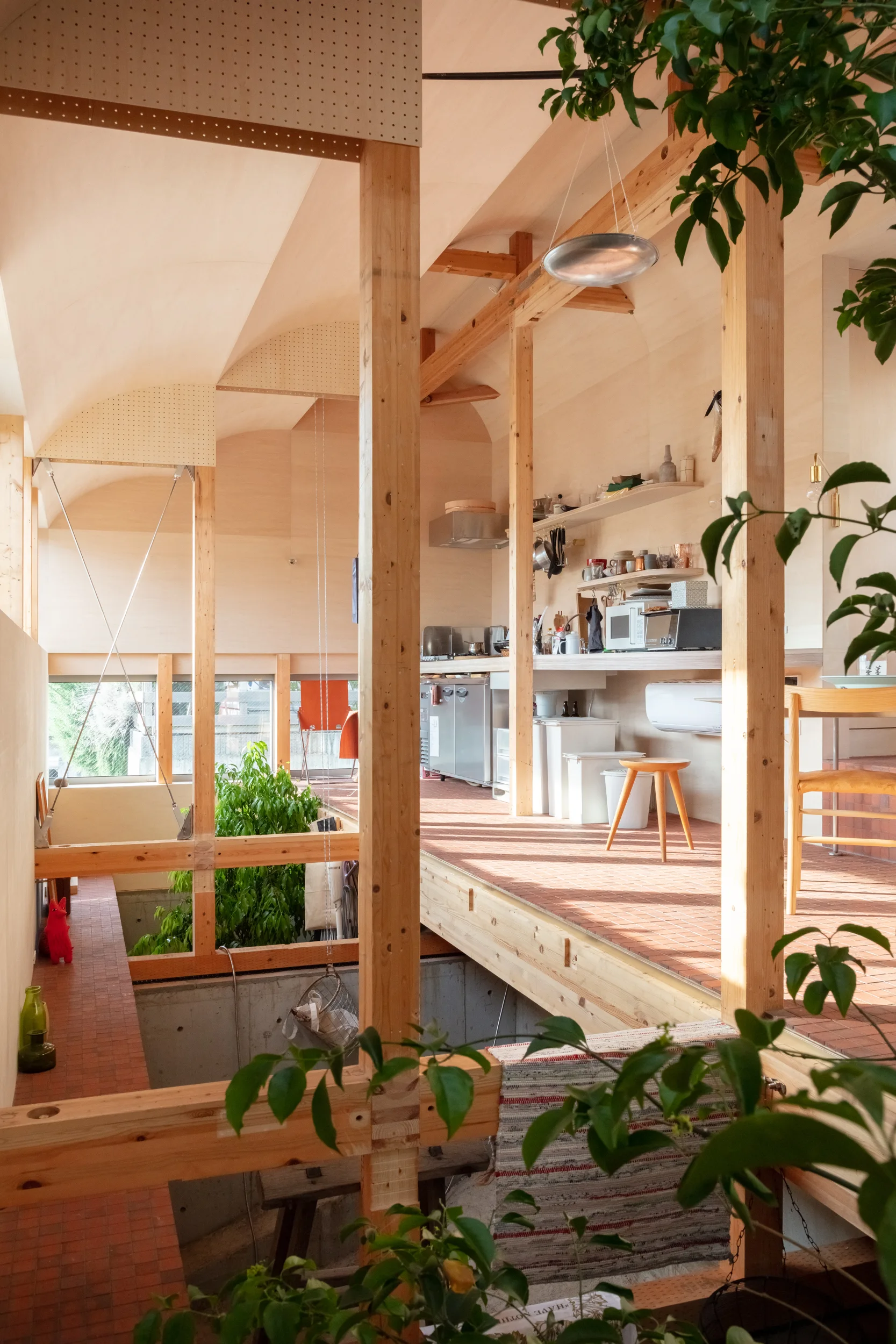 A minimalist residential kitchen featuring exposed timber posts, a curved light wood ceiling, and red terracotta floor tiles.