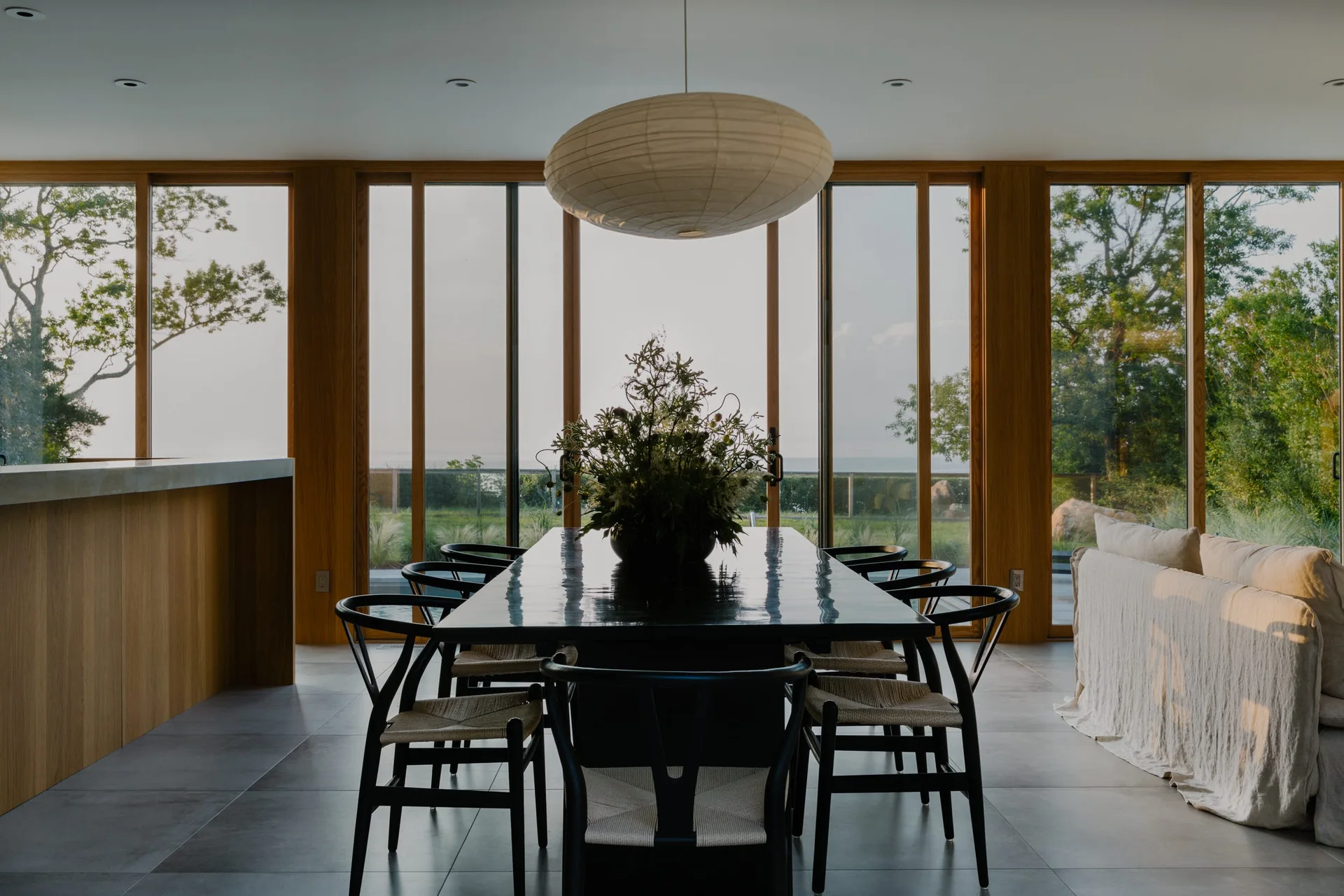 A modern dining room featuring a long black table, black chairs, and a large paper pendant lamp in front of wood-framed windows.