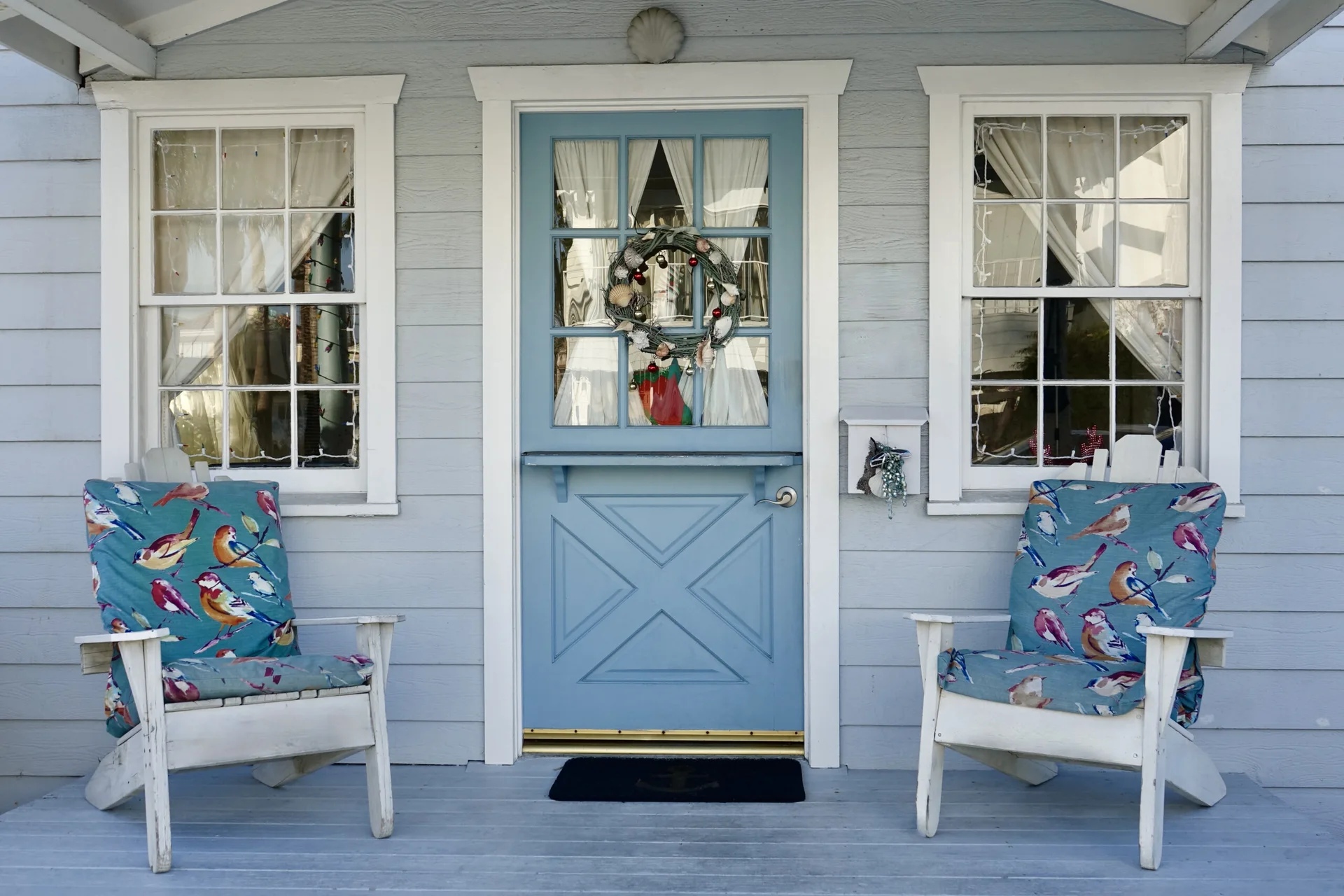 A light blue cottage porch features a matching Dutch door with a wreath, flanked by two white wooden chairs with bird-patterned blue cushions.