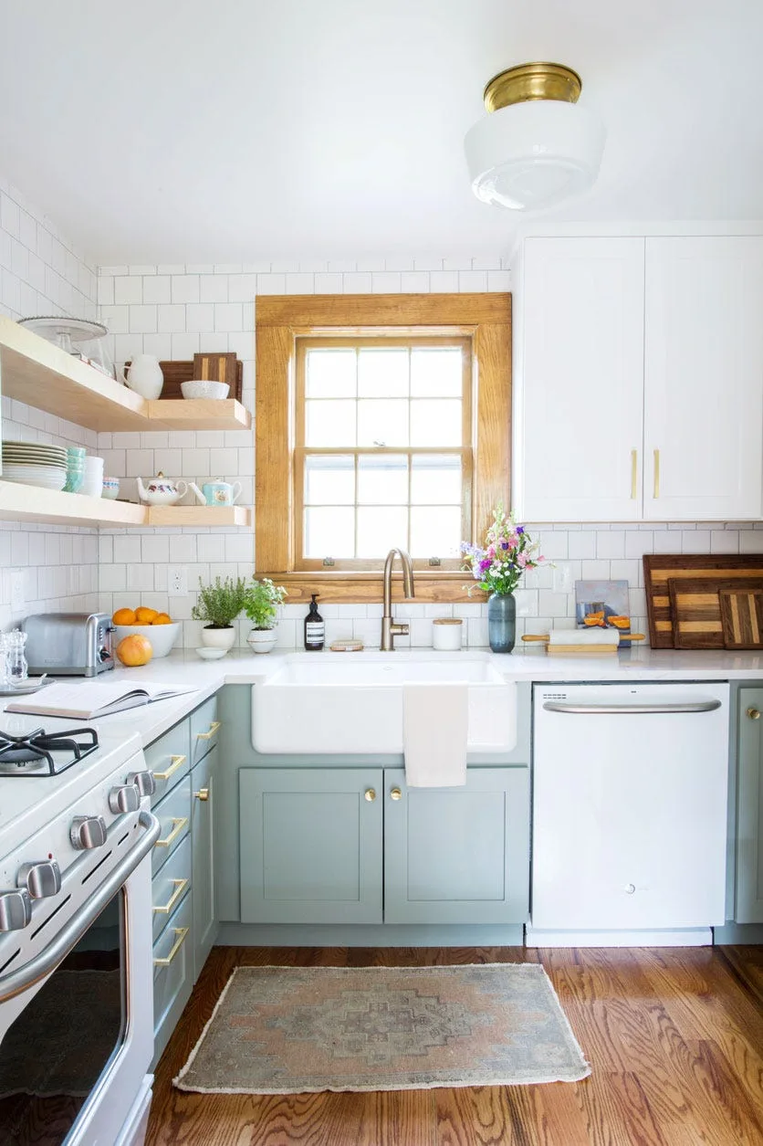 A kitchen with sage green base cabinets, a white farmhouse sink, floating wood shelves, and a natural wood-trimmed window over white subway tile.