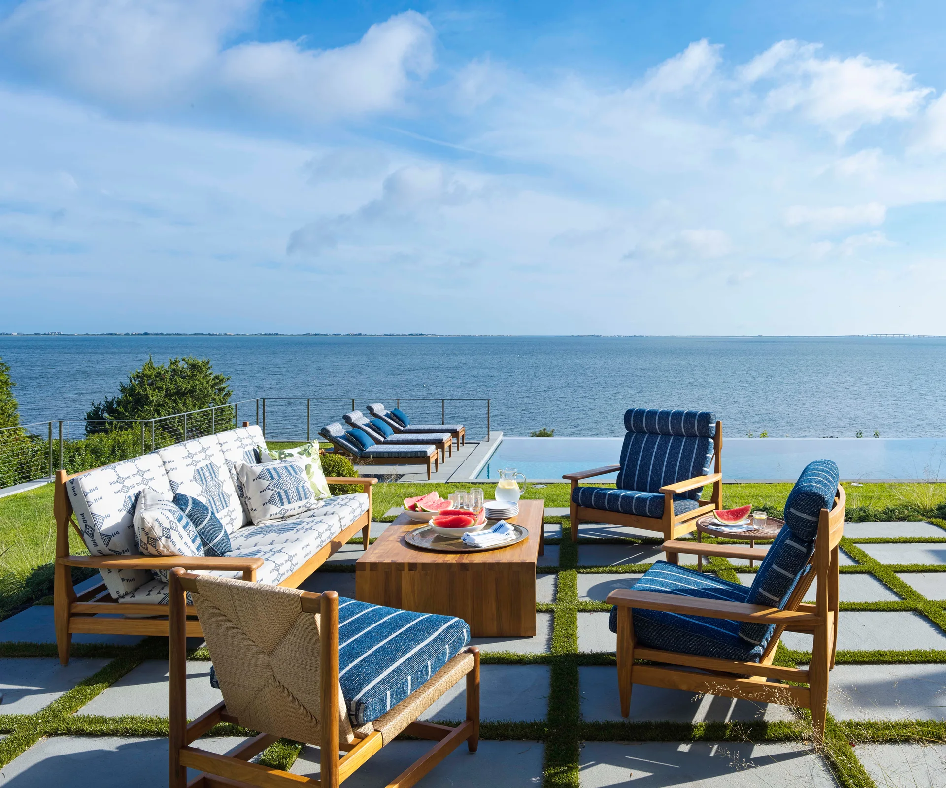 Wooden patio furniture with blue and white patterned cushions sits on stone grid pavers overlooking an infinity pool and the ocean.