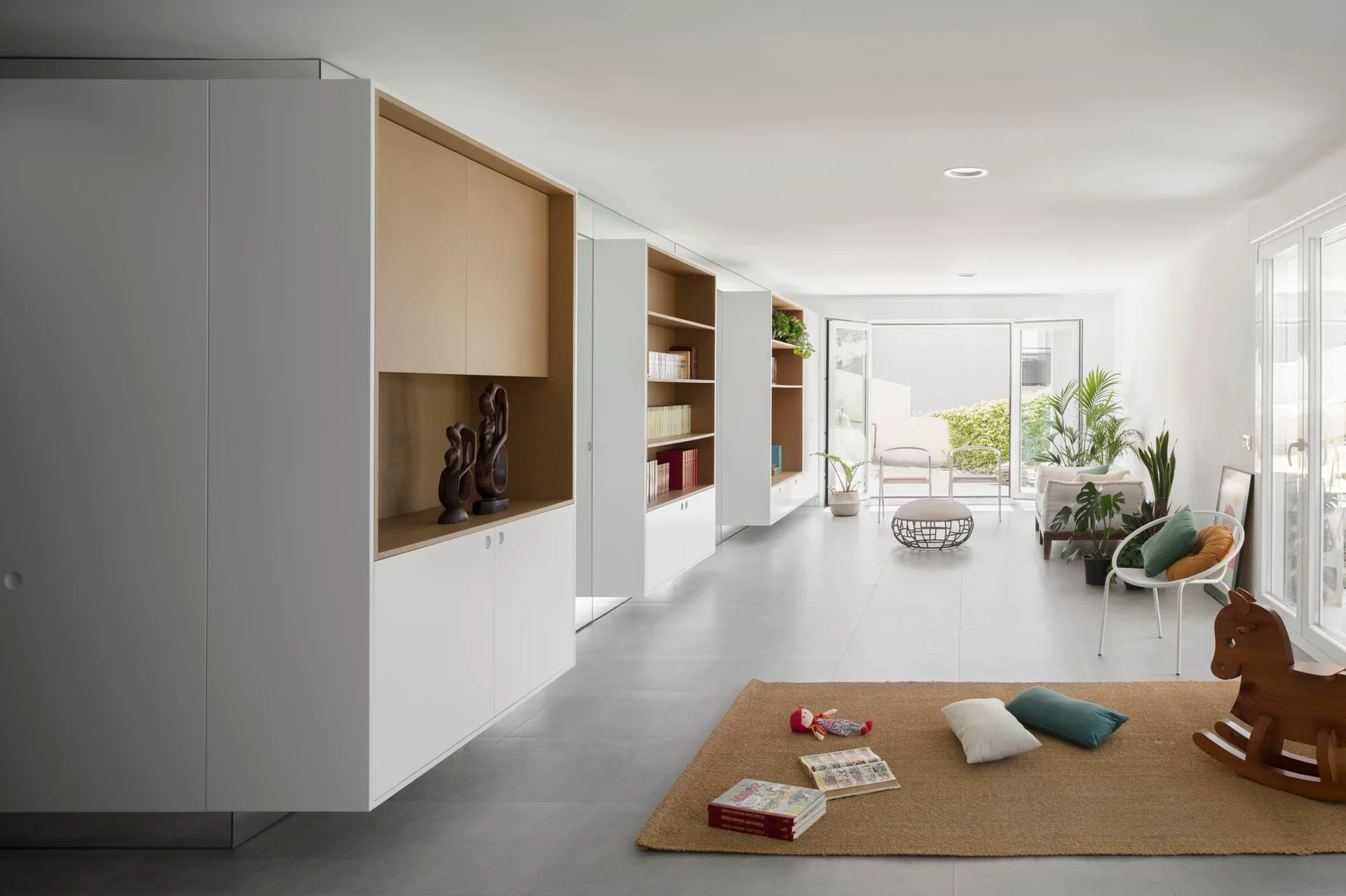 A spacious minimalist room featuring grey tile flooring, white built-in cabinets with wood-toned shelves, a tan area rug, and floor-to-ceiling windows.