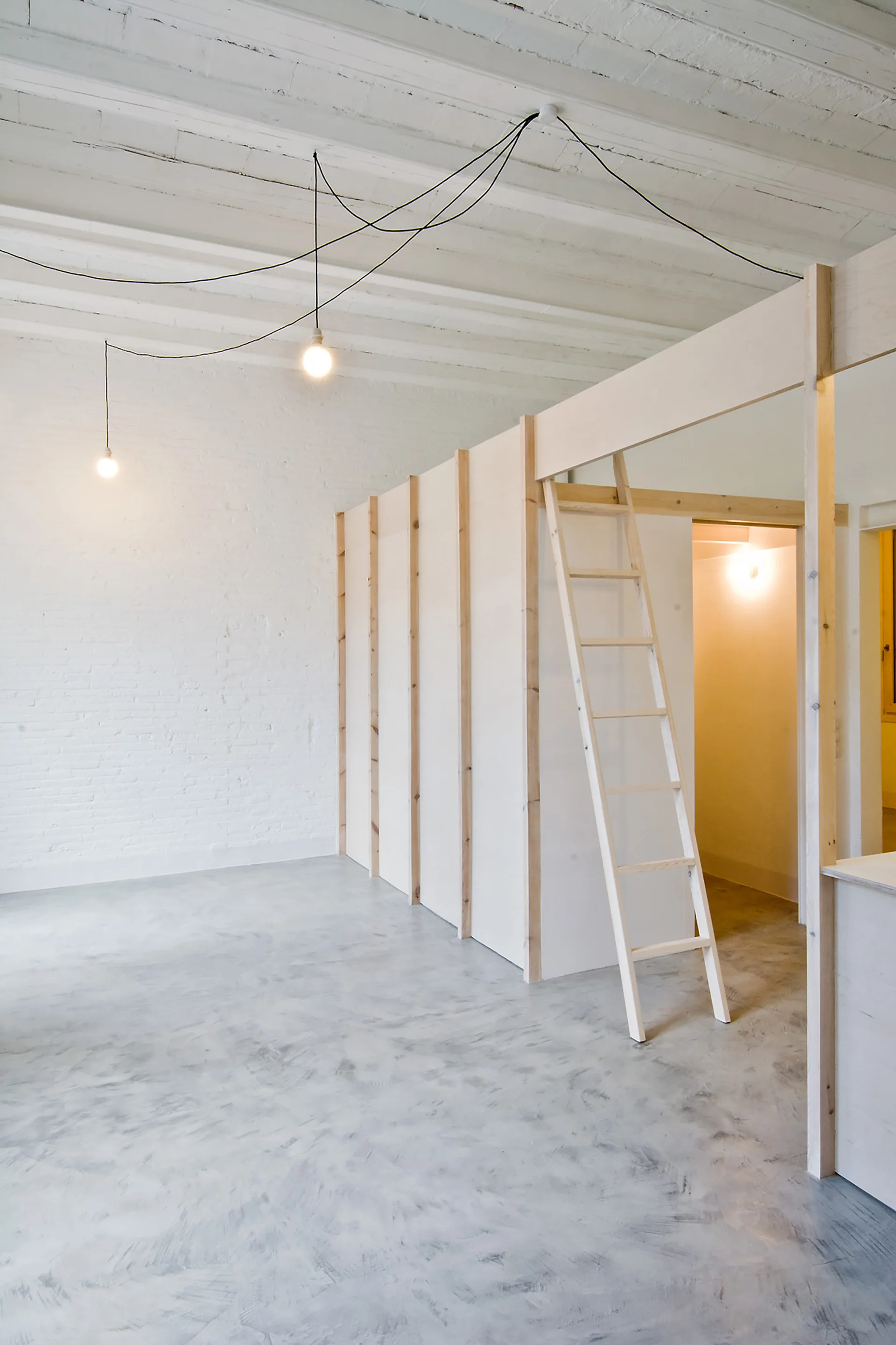 A minimalist loft space features a white painted brick wall, mottled grey concrete floors, and a light wood partition wall with an integrated ladder.