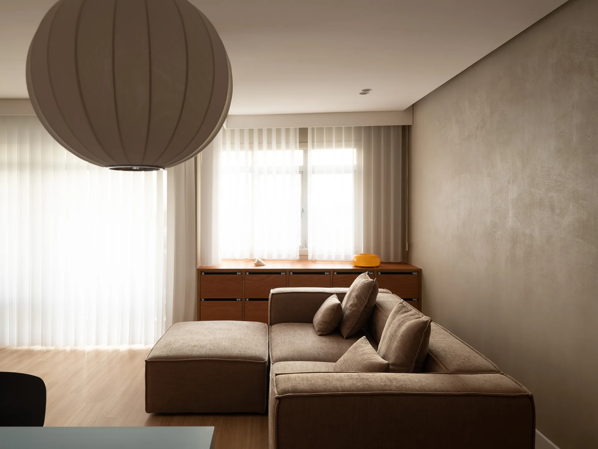 A minimalist living room features a tan fabric sectional sofa, a low wooden storage console under a window with sheer curtains, and a large round pendant light.