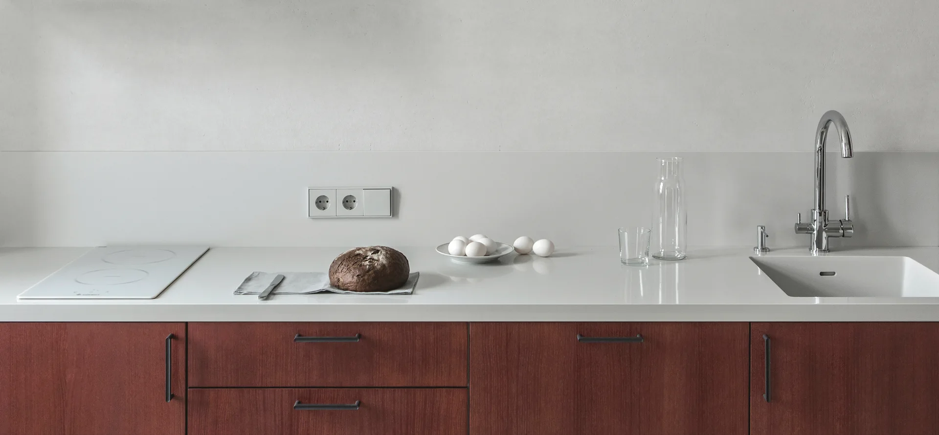 A minimalist kitchen counter features dark reddish-brown wooden cabinets, a seamless white countertop, a white induction cooktop, and a chrome faucet.