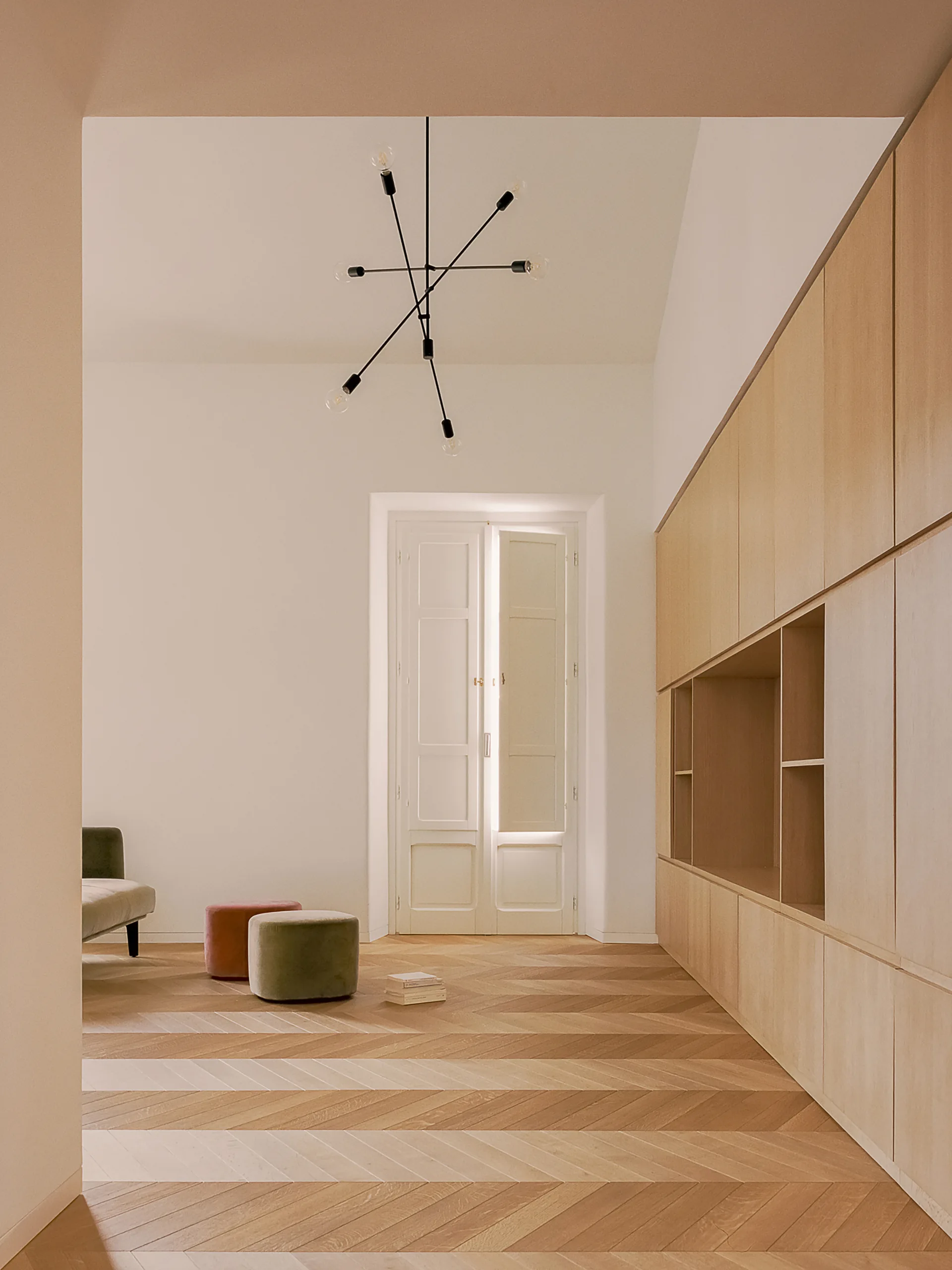 A minimalist room features light wood herringbone flooring, a large built-in wooden cabinet wall, and two small velvet ottomans.
