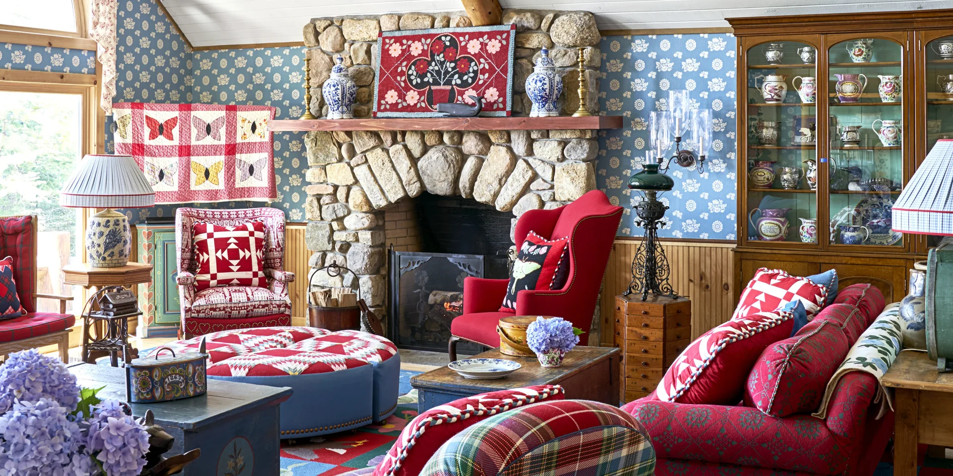 A cozy living room with a rustic stone fireplace, blue floral wallpaper, red quilted furniture, and a large wooden display cabinet.